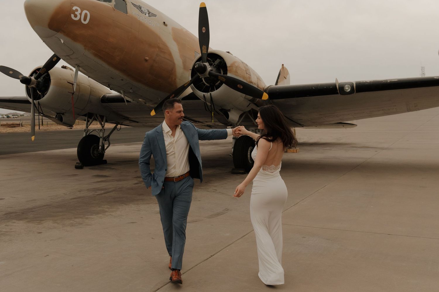 Couple dancing on tarmac in front of vintage WWII airplane, bride in white gown, groom in blue suit.