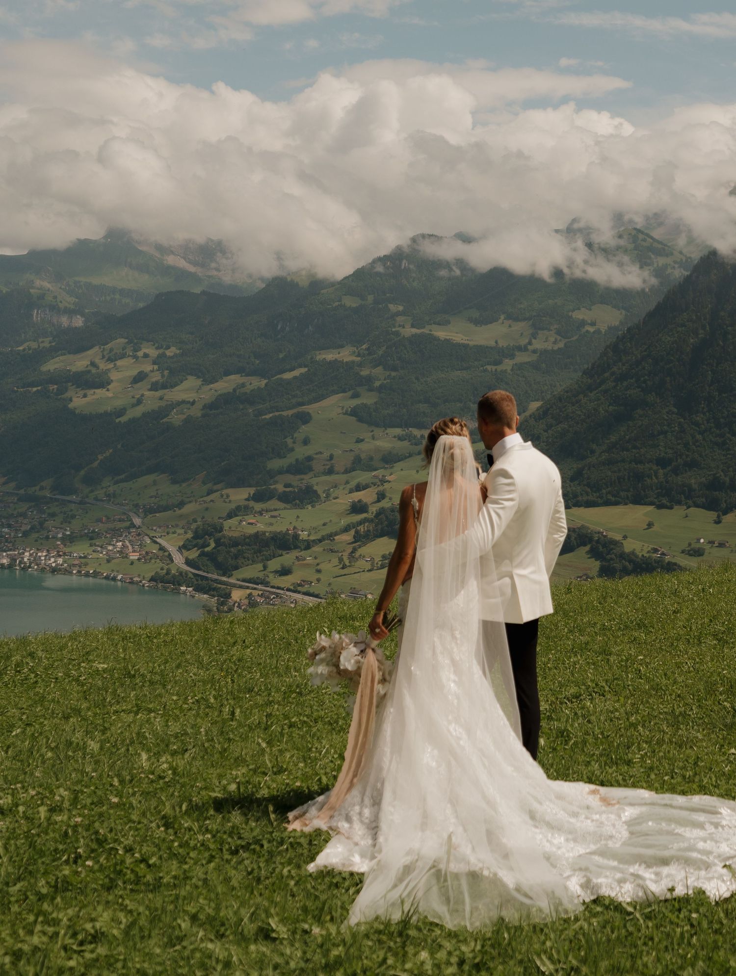 Bride and groom standing back to camera overlooking stunning Swiss alpine valley and lake.