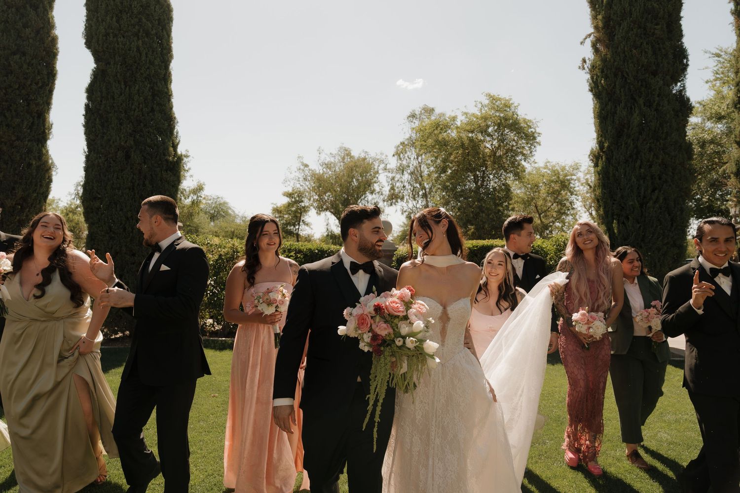 Bride and groom walk with wedding party through lush garden, bride holding pink and white floral bouquet.