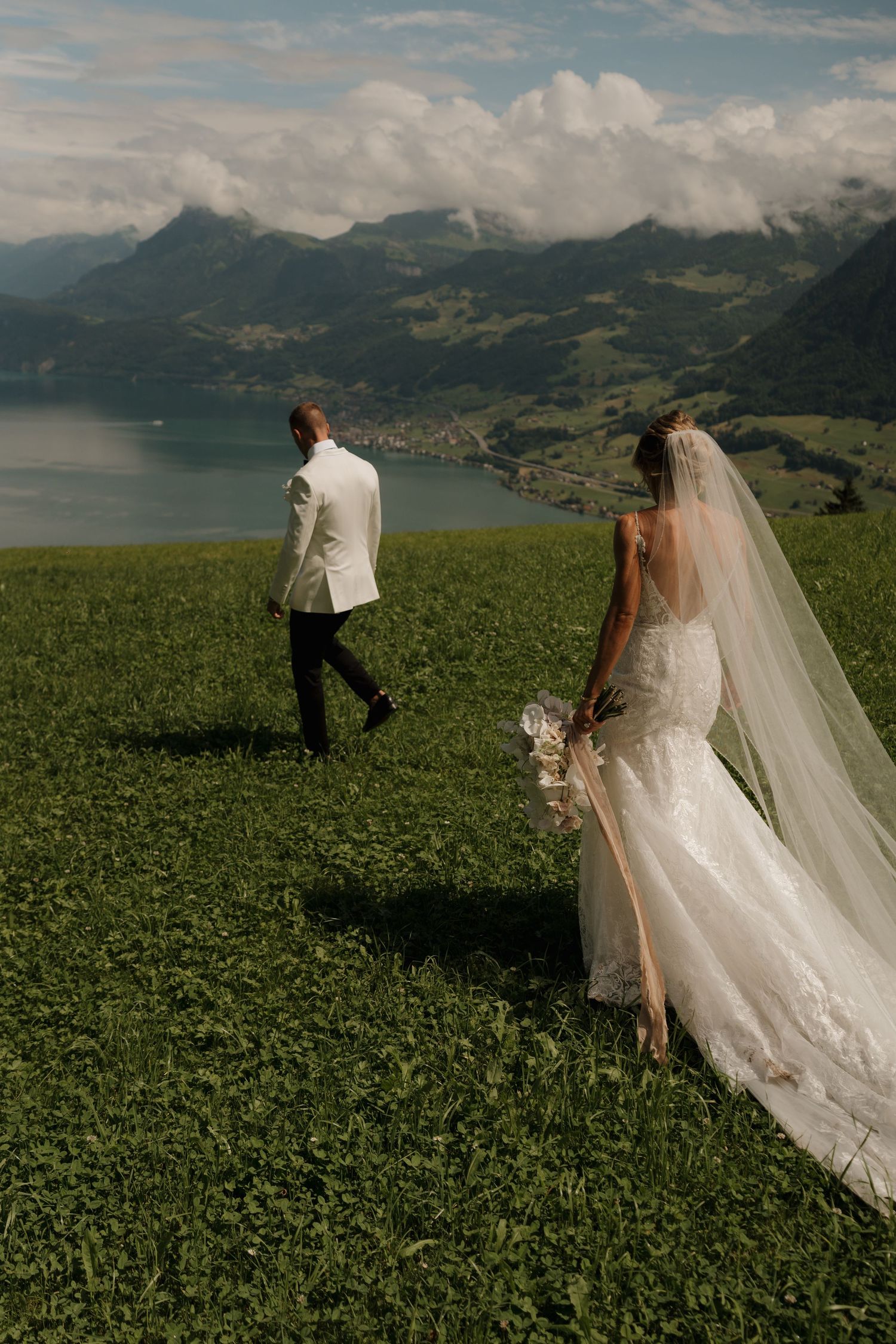 Bride and groom walk through lush green alpine meadow with stunning lake and mountain views in Switzerland.