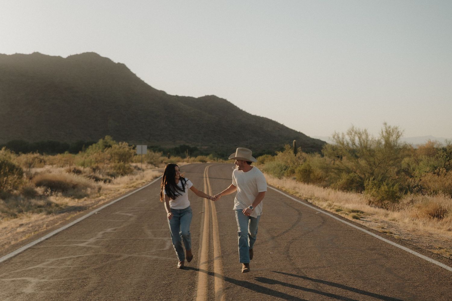 Couple holds hands walking apart on empty desert road with mountain landscape at sunset.