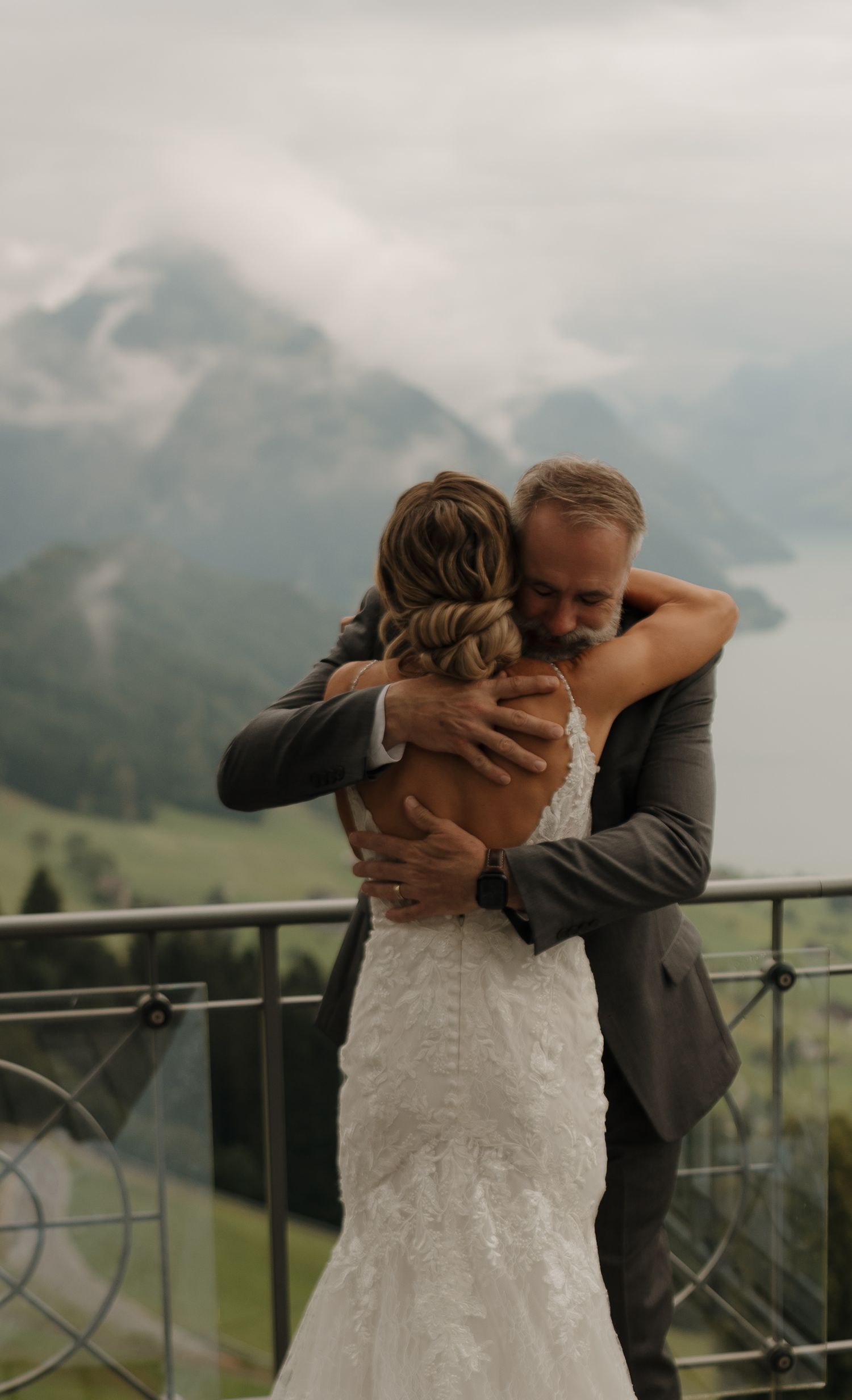 Emotional embrace between bride in lace gown and wedding guest on a balcony with misty mountain lake backdrop.