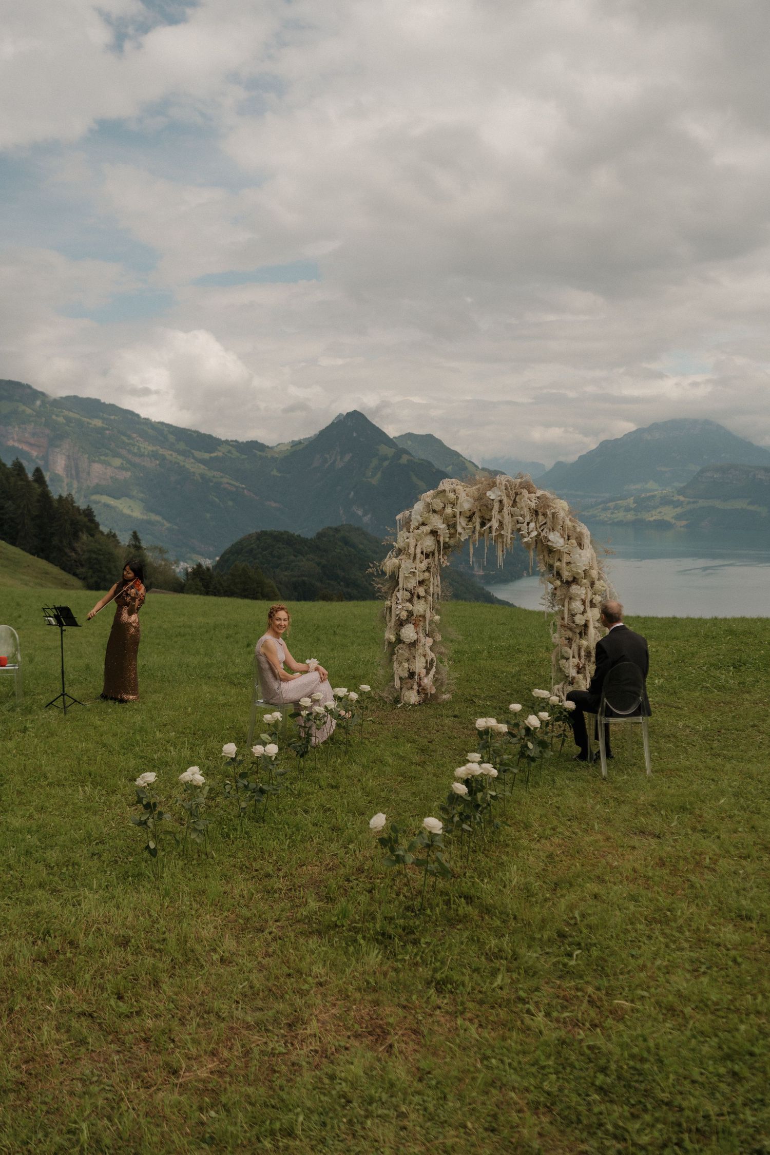Outdoor wedding ceremony setup with a floral arch on a green meadow overlooking a scenic alpine lake.