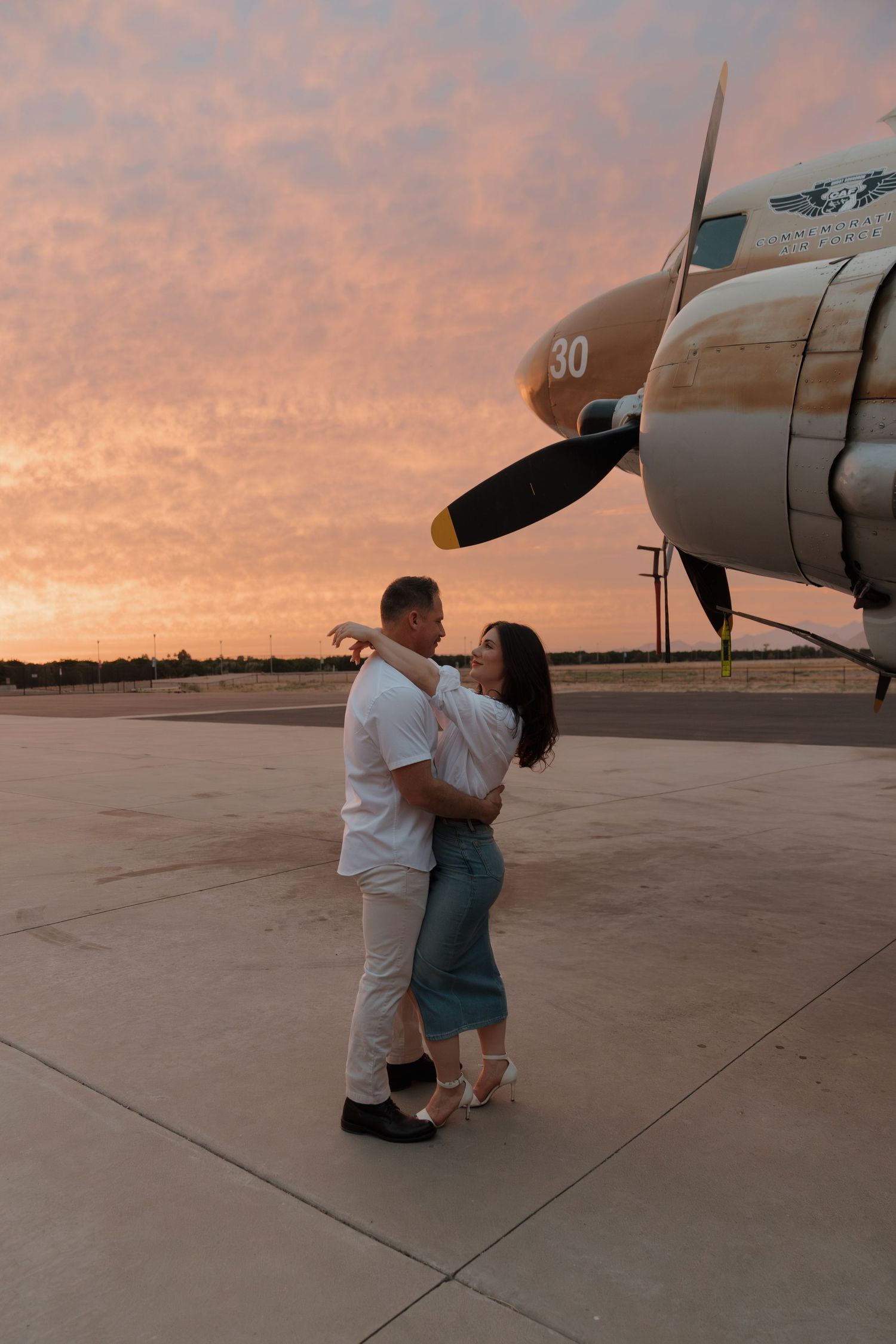 Couple embracing romantically on airport tarmac beside vintage propeller plane at stunning pink and orange sunset.