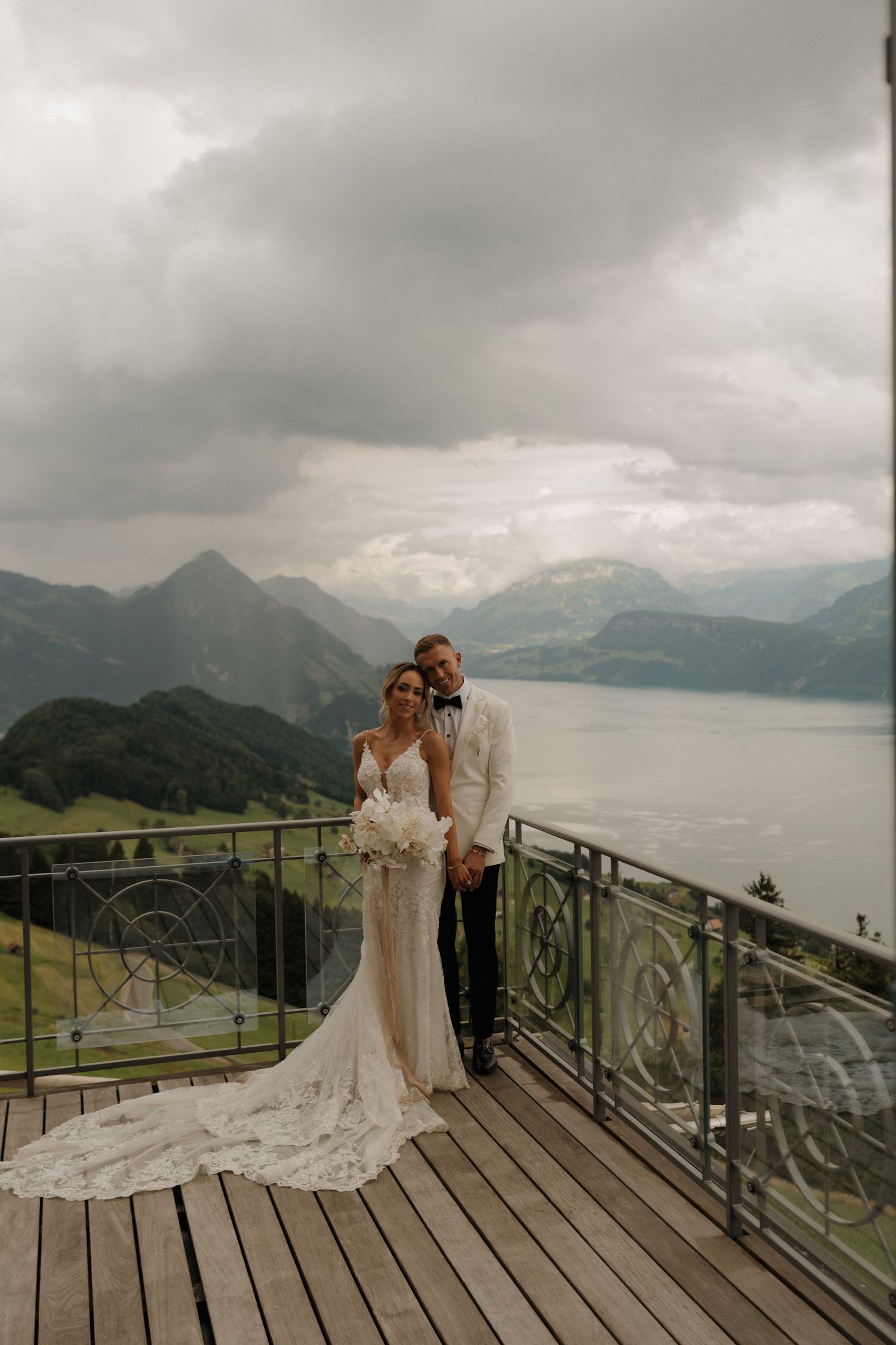 Bride and groom posing on wooden terrace with stunning alpine lake and mountain views behind them.