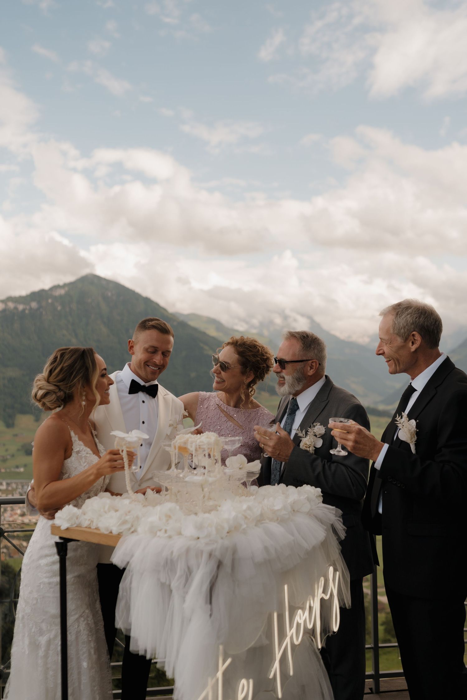 Bride, groom, and family toasting with champagne glasses on a scenic mountain terrace after wedding.