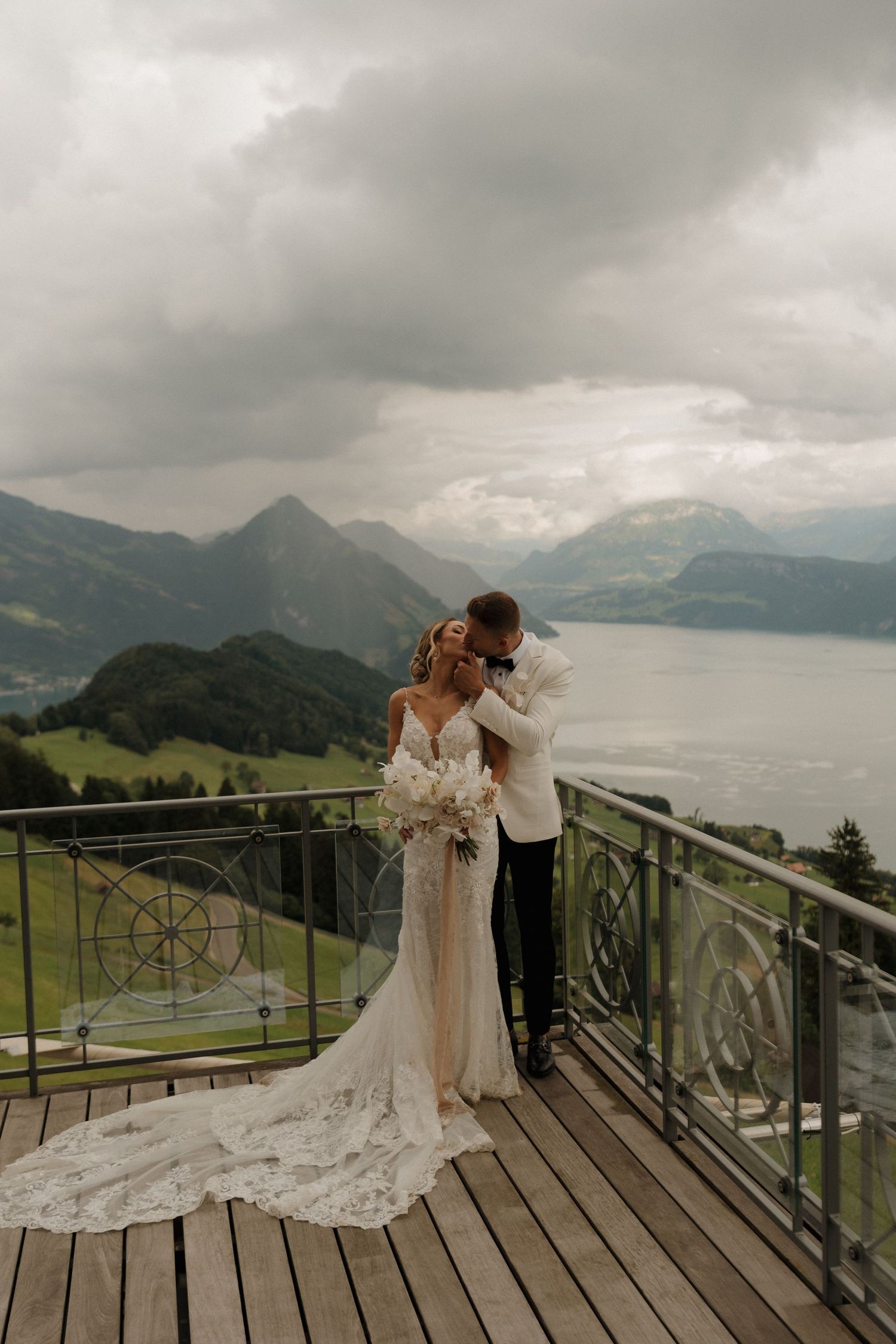 Elegant couple embraces on wooden terrace overlooking dramatic Swiss Alps lake under moody cloudy skies.