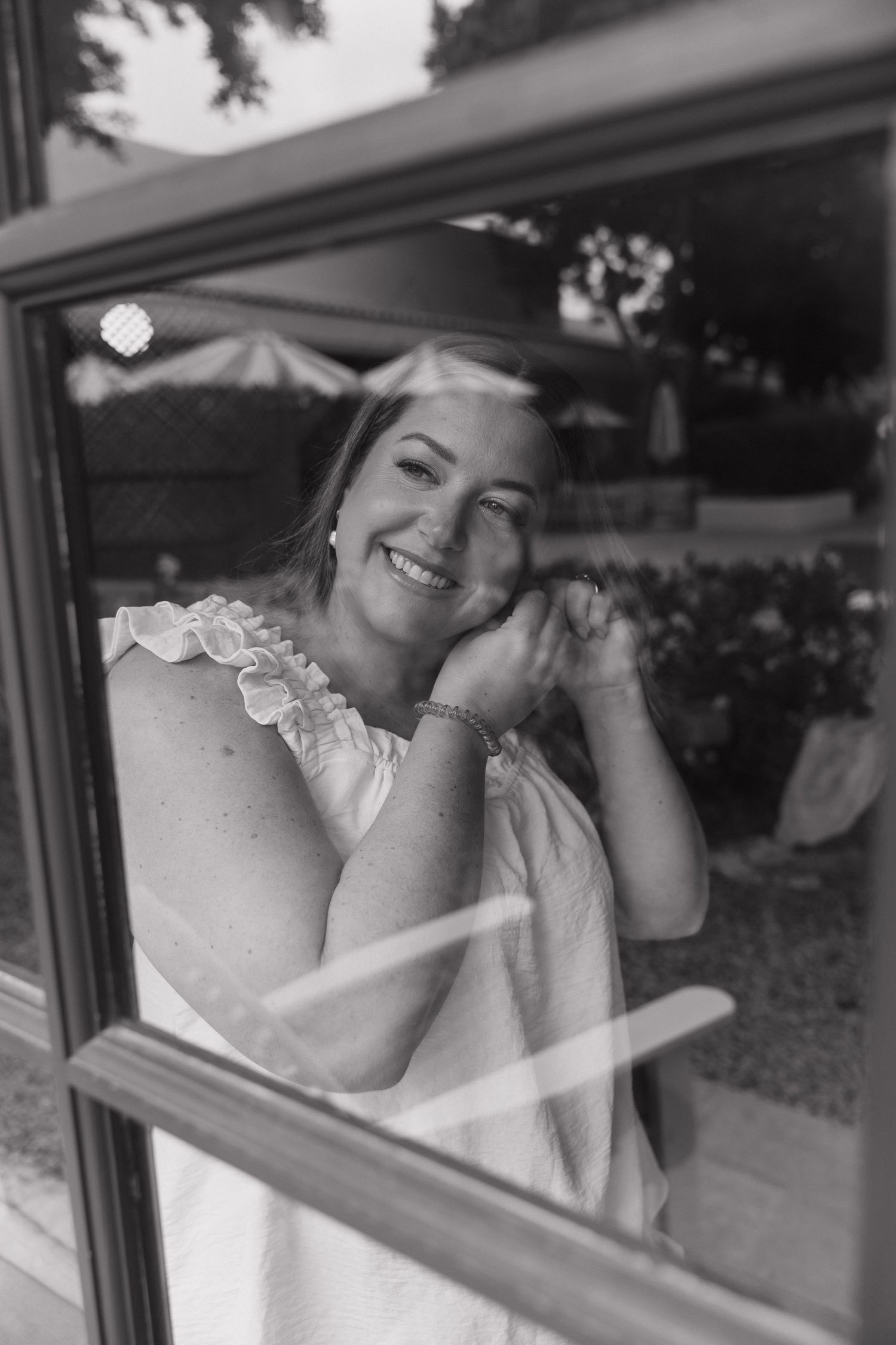 Black and white photo of smiling bride in white dress seen through a window reflection.