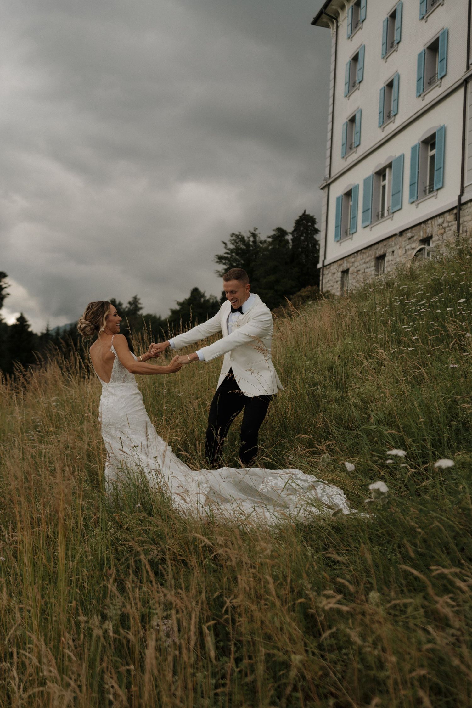 Bride and groom dancing joyfully in tall grass beside historic building under dramatic sky.