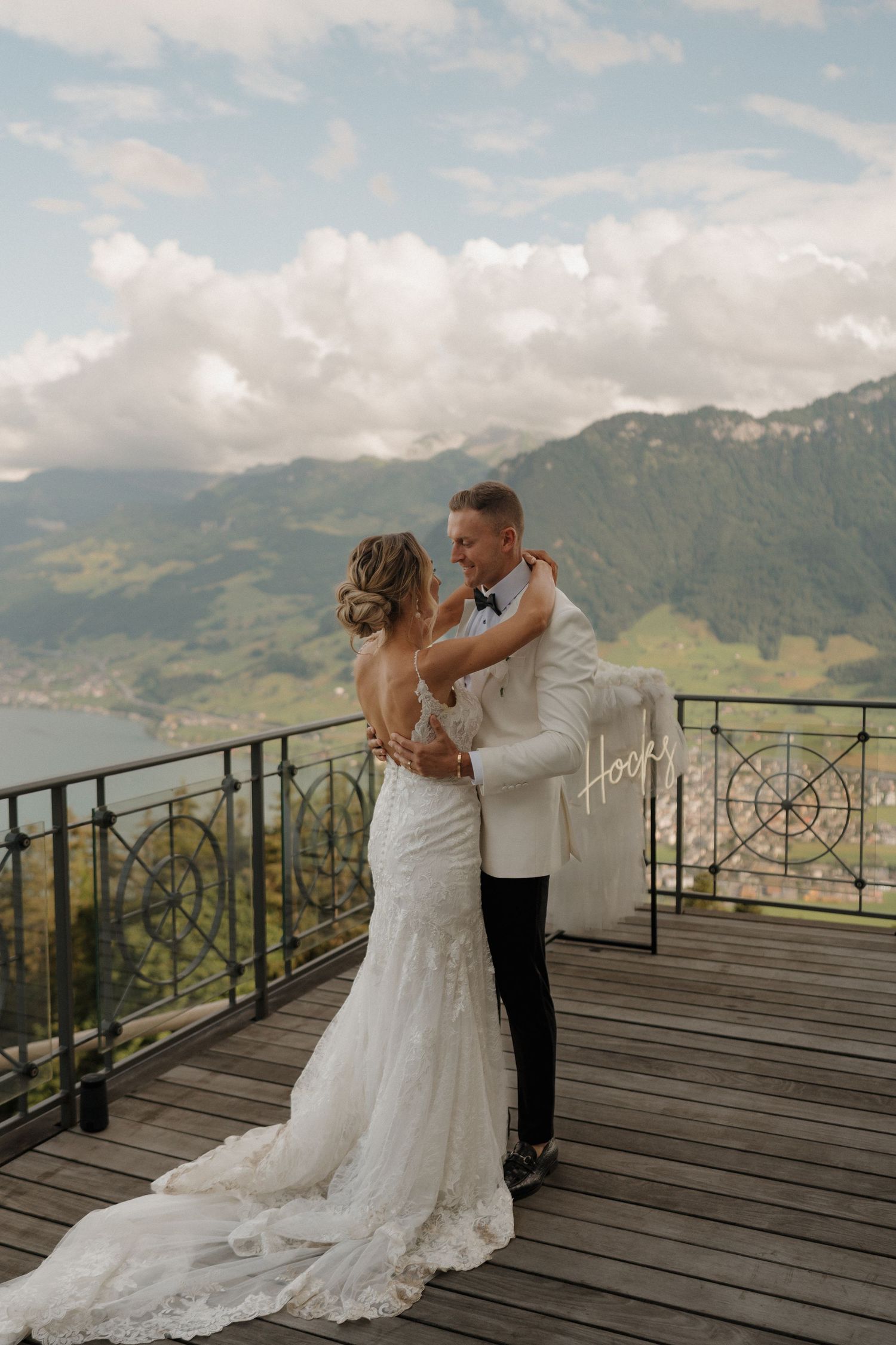 Bride and groom embrace on scenic mountain balcony, with stunning Alpine lake backdrop on wedding day.