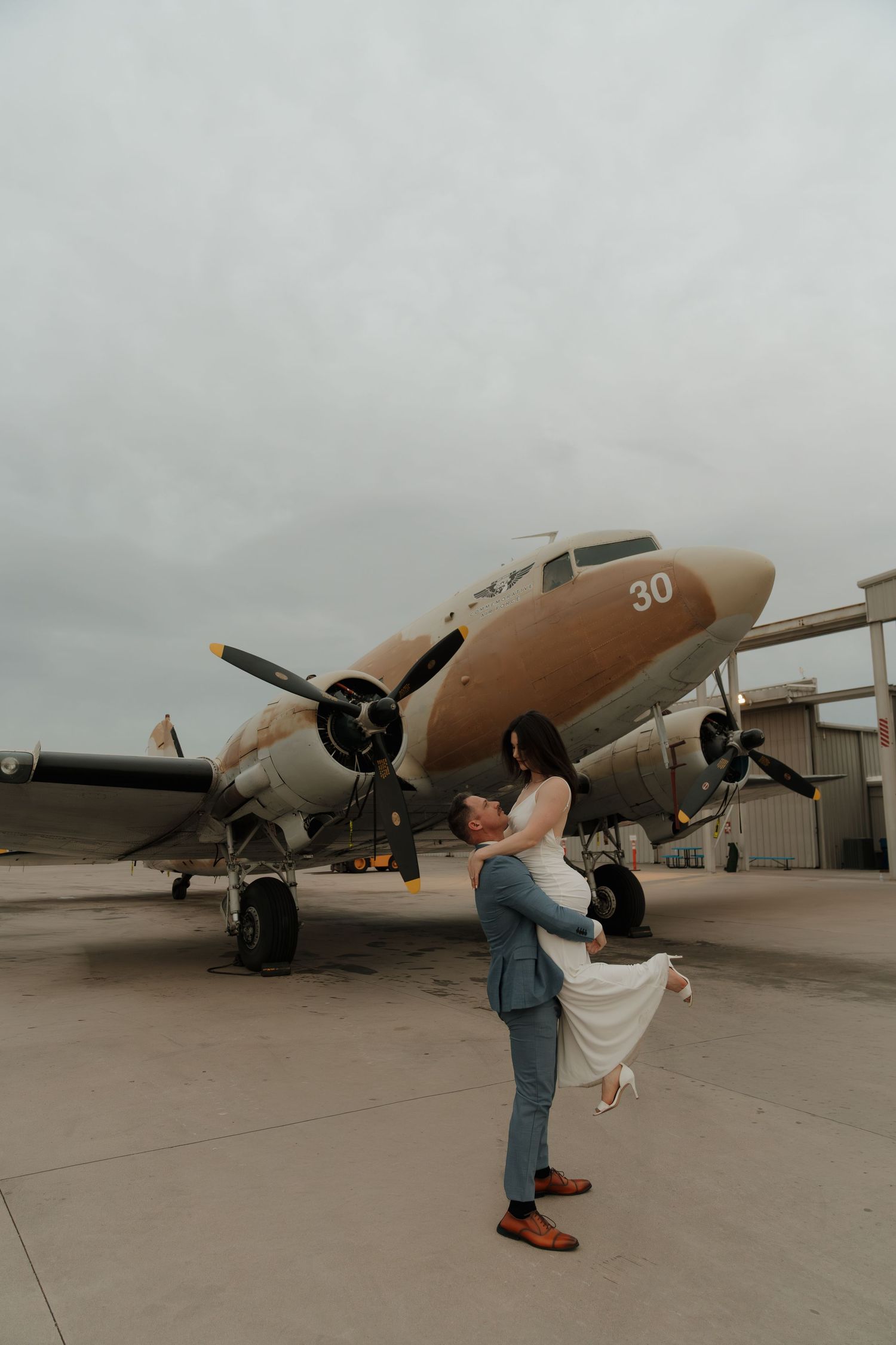Groom lifting bride in front of vintage propeller plane numbered 30 on airport tarmac under overcast skies.