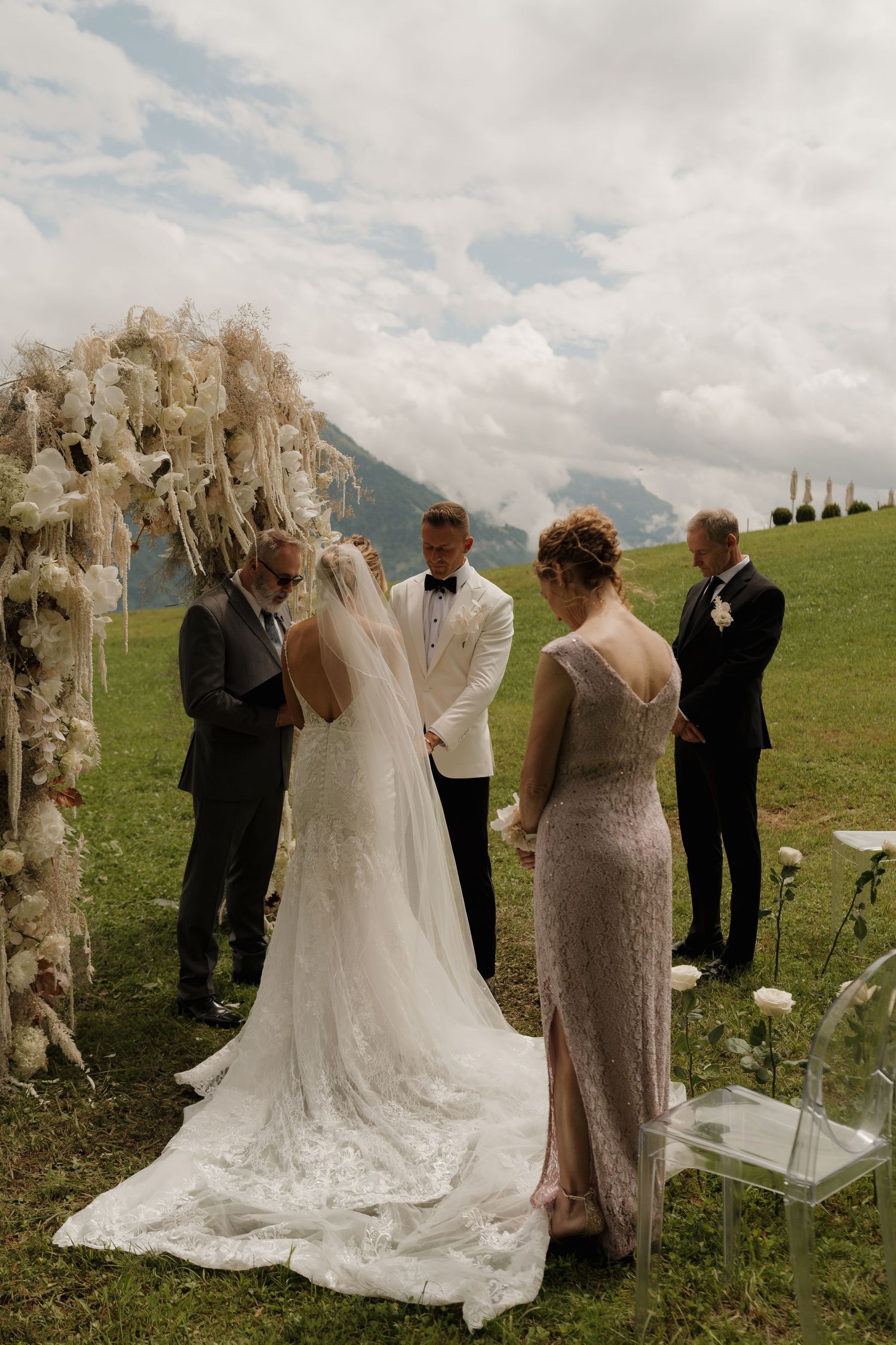 Bride and groom exchange vows under a stunning white floral arch during an outdoor alpine wedding ceremony.