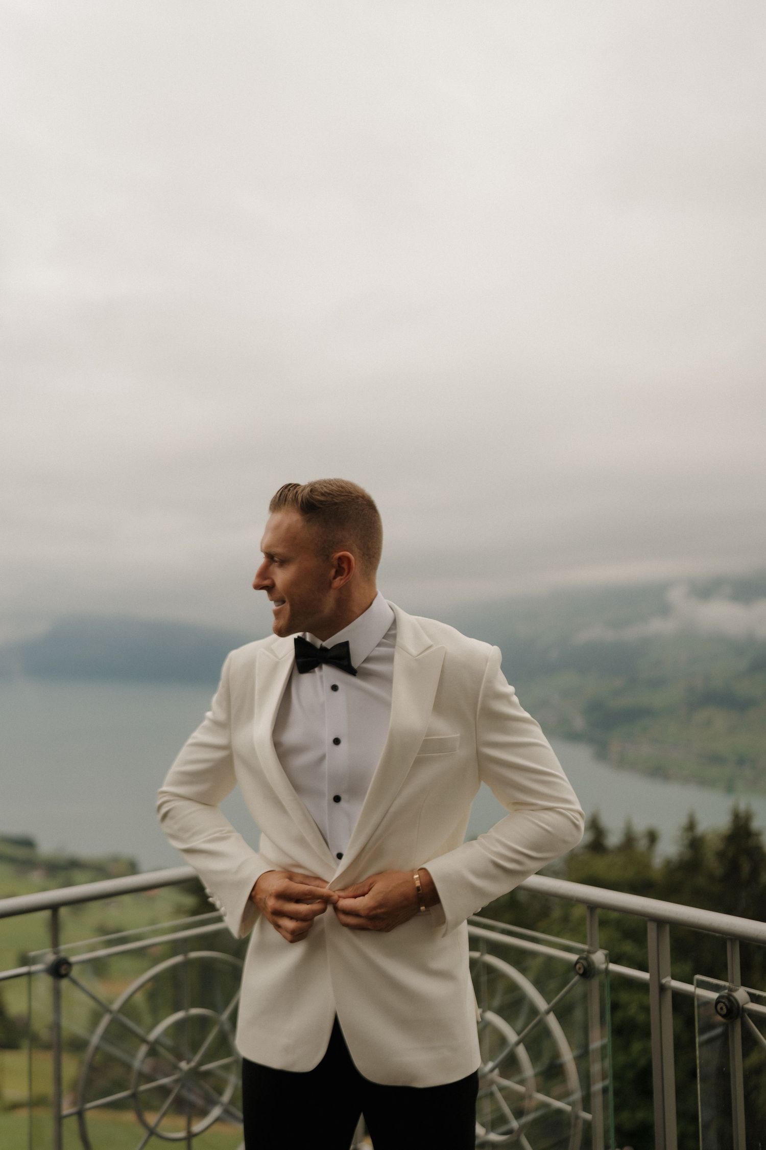 Dapper groom in white tuxedo jacket and black bow tie standing on a balcony overlooking a misty Alpine lake.