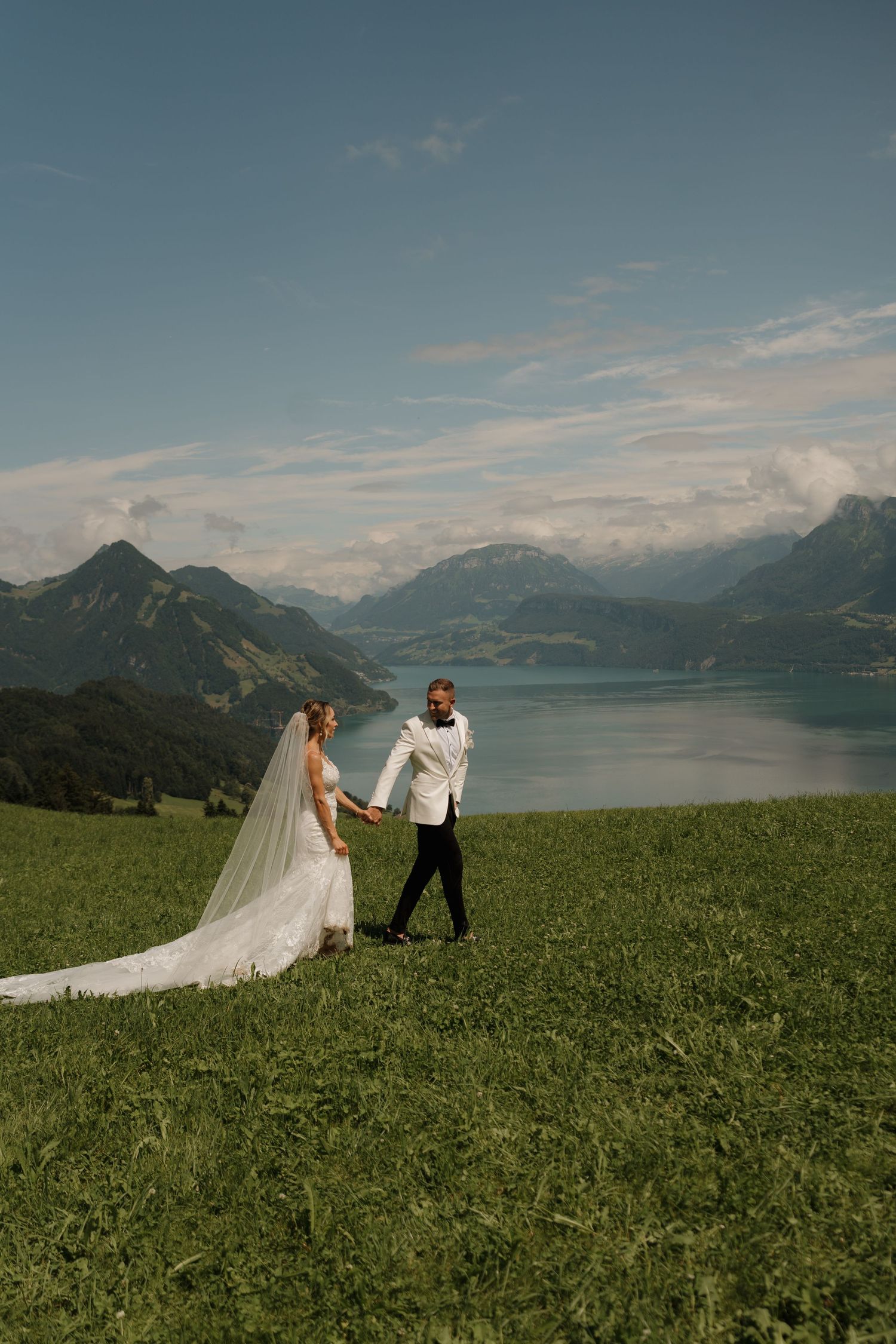 Bride and groom hold hands on green hillside with breathtaking Swiss Alps lake panorama behind them.