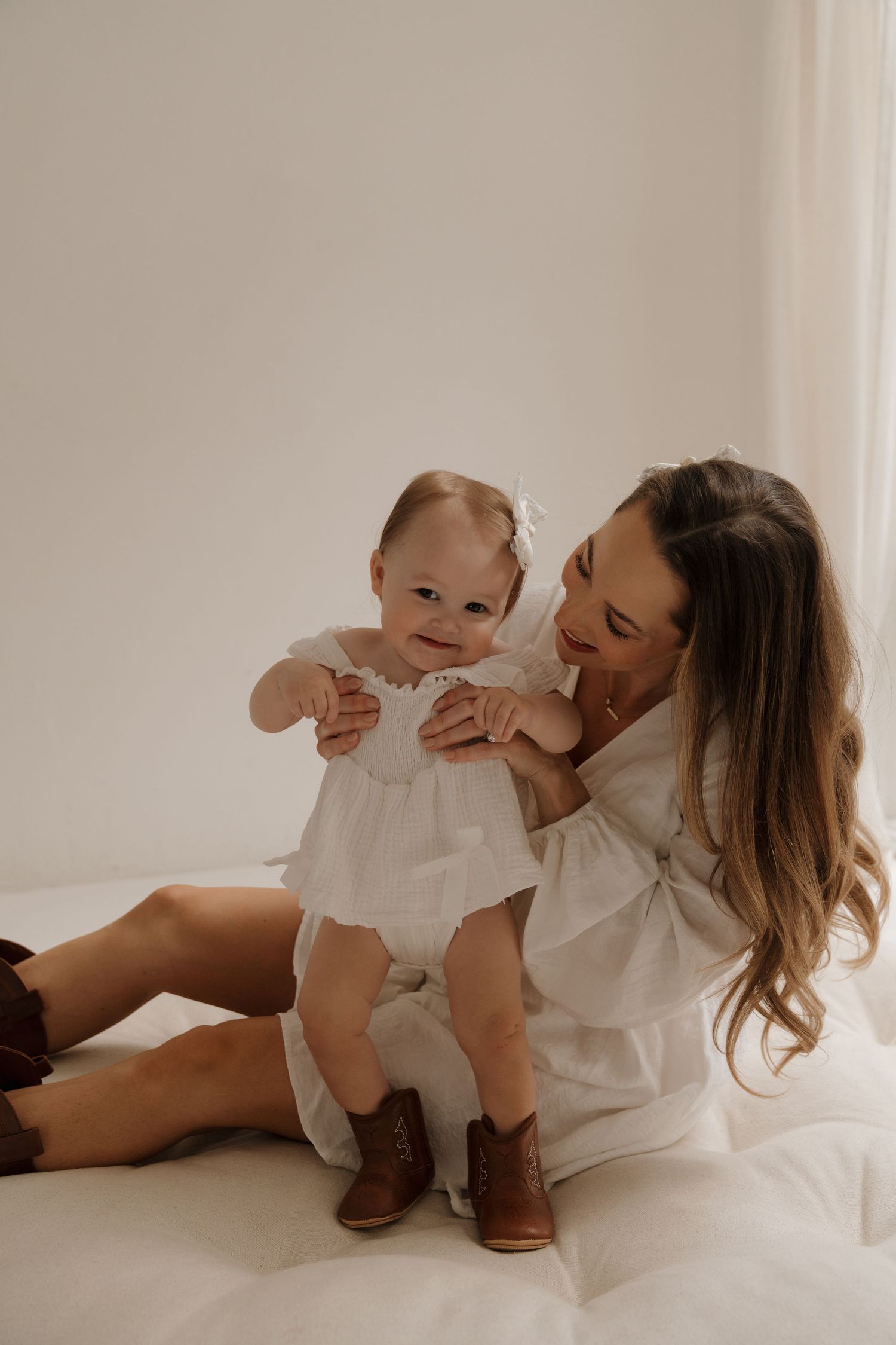 Mother in white dress sitting on bed helping baby girl in white dress stand up.