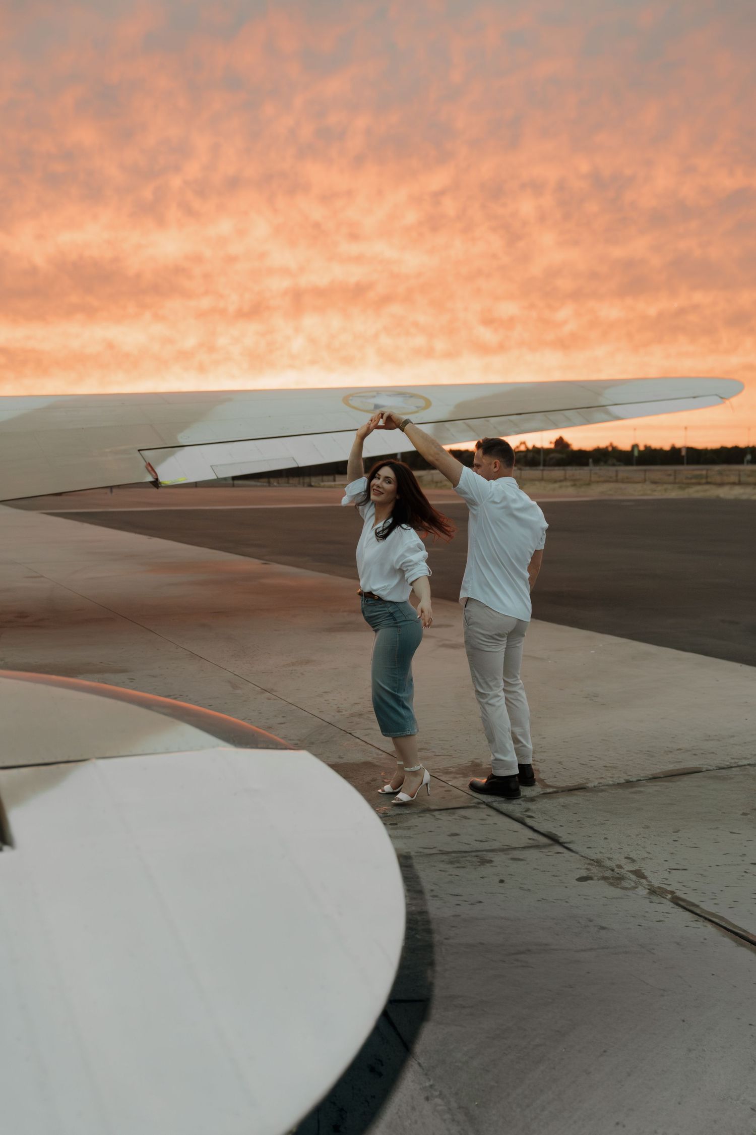 Couple dancing on airport tarmac near airplane wing at sunset with vibrant orange sky backdrop.