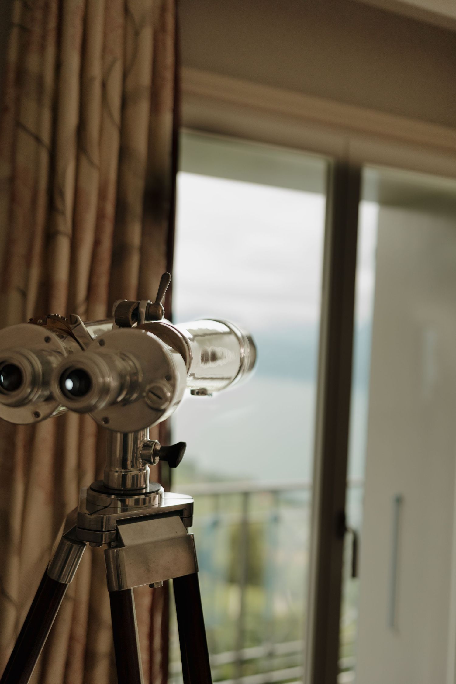 Vintage binoculars on tripod stand indoors with mountain view through window and beige curtains.