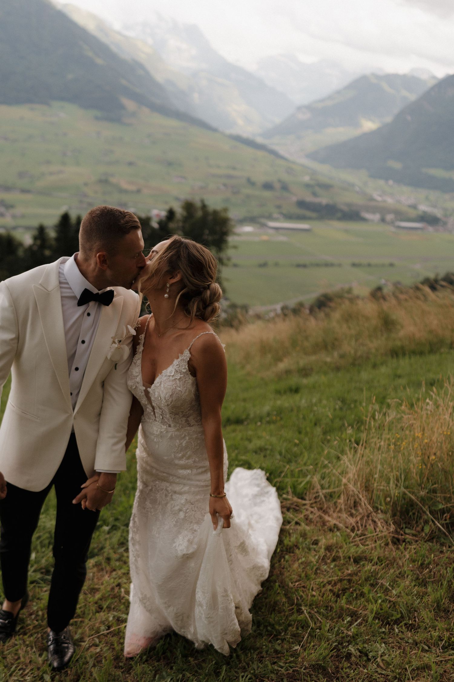Bride and groom share a kiss on a scenic alpine hillside, groom in white tuxedo jacket.