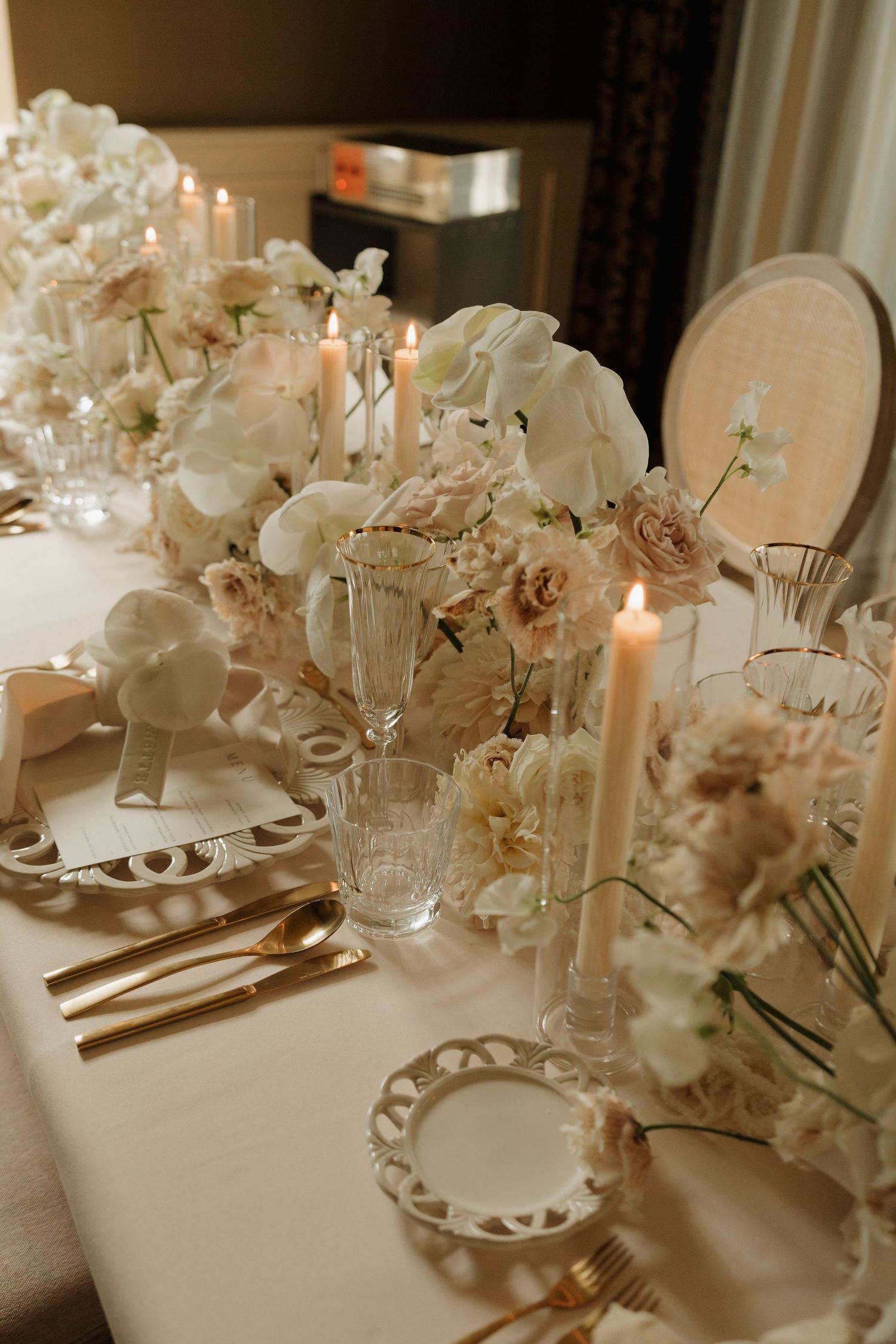 Elegant wedding reception table with white orchids, blush roses, lit candles, gold cutlery, and ornate white plates.