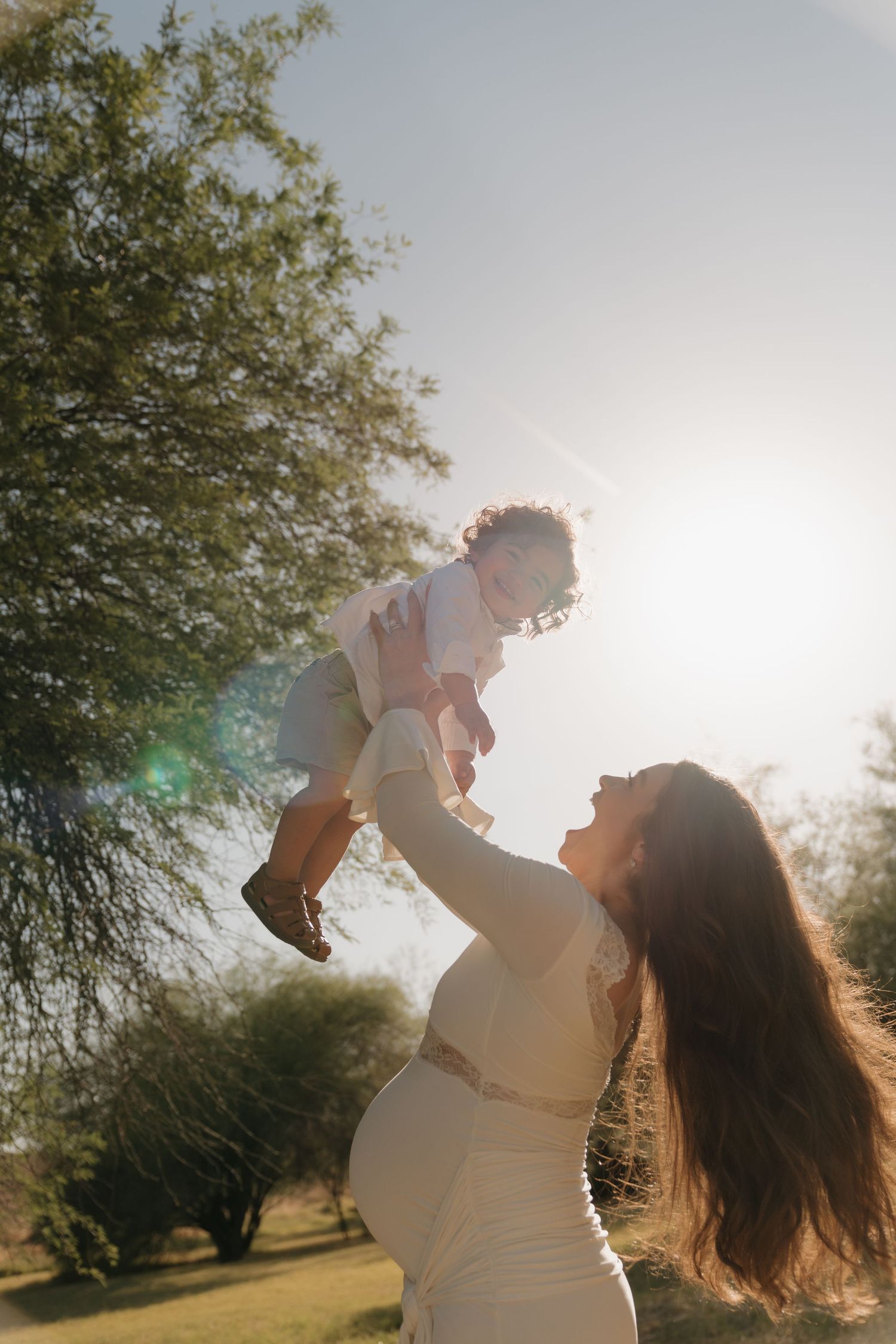 Pregnant woman in white dress lifting toddler against sunny sky with trees in background.