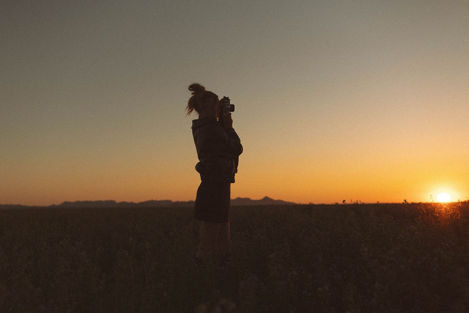 Silhouette of a woman embracing in a field during golden hour sunset with mountains in the background.