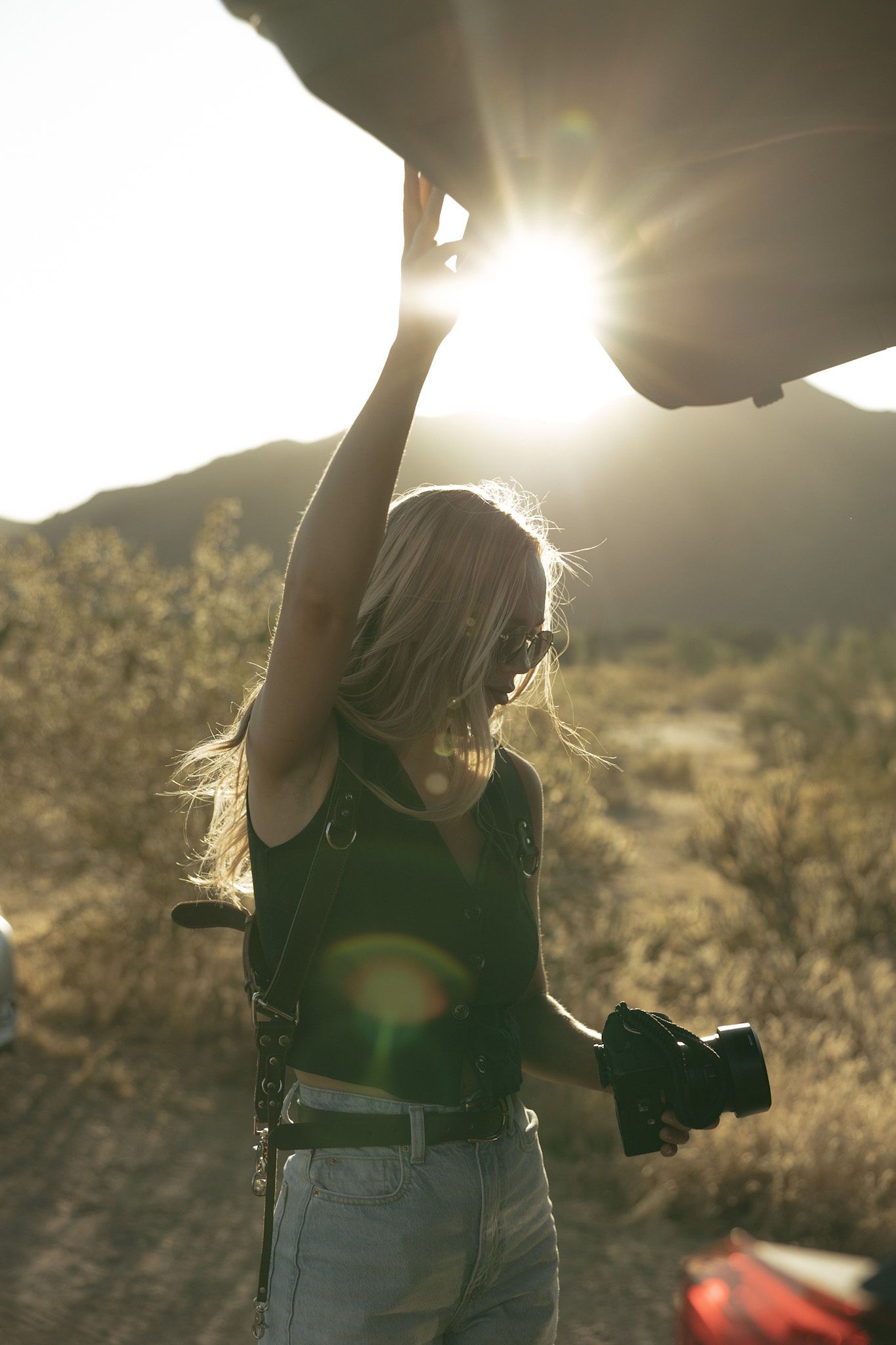 Blonde woman holding open a car trunk with a DSLR camera in hand, backlit by golden sunlight in a desert setting.