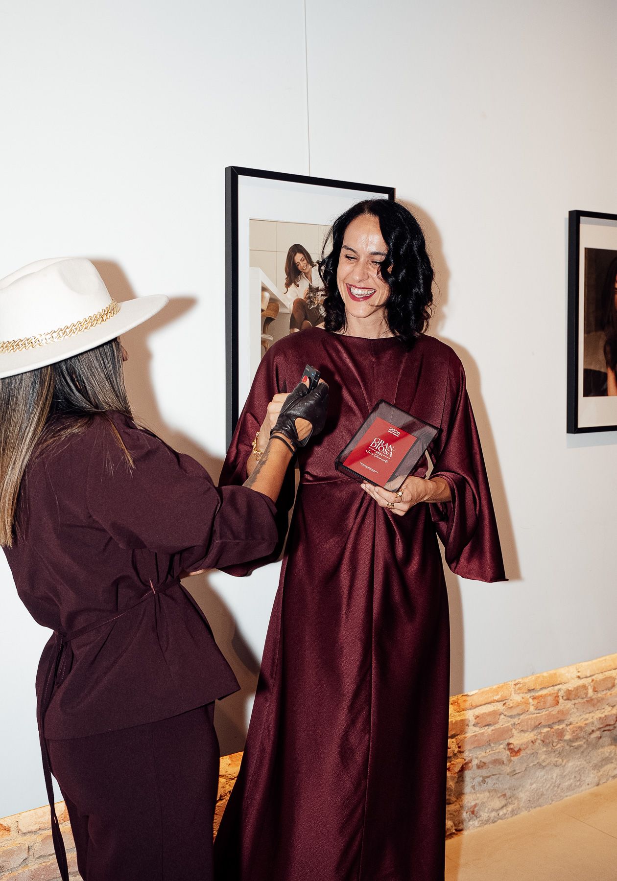 Two women in burgundy outfits at an art gallery, one holding a red book while being photographed.