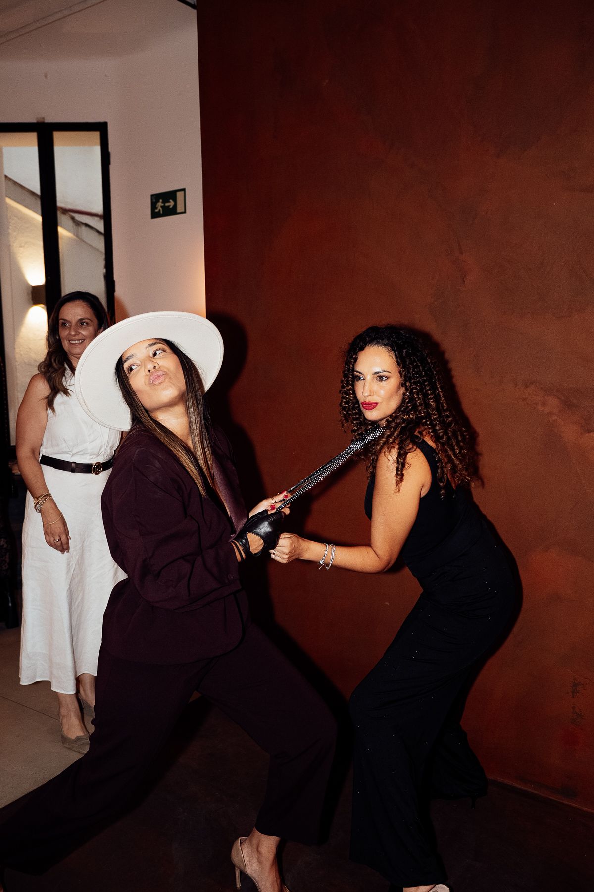 Two women playfully pose with a sword while a third woman watches and smiles in an elegant indoor setting.