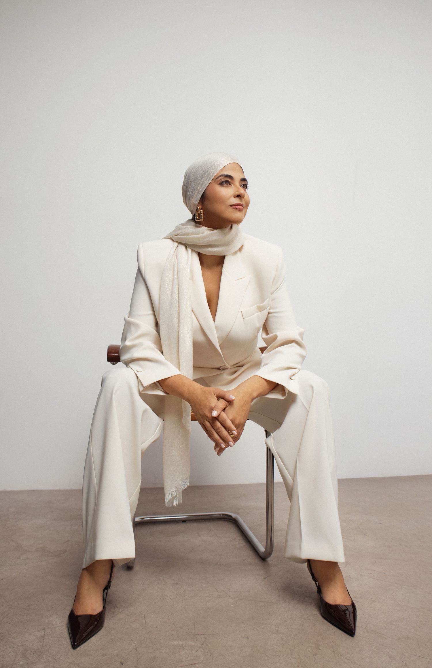 Elegant woman in cream white suit and hijab seated on chair, posing in minimalist studio setting.
