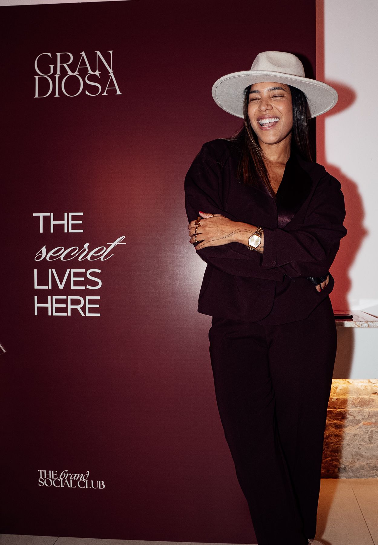 Woman in white hat and dark suit laughing at Gran Diosa event with 'The Secret Lives Here' backdrop.