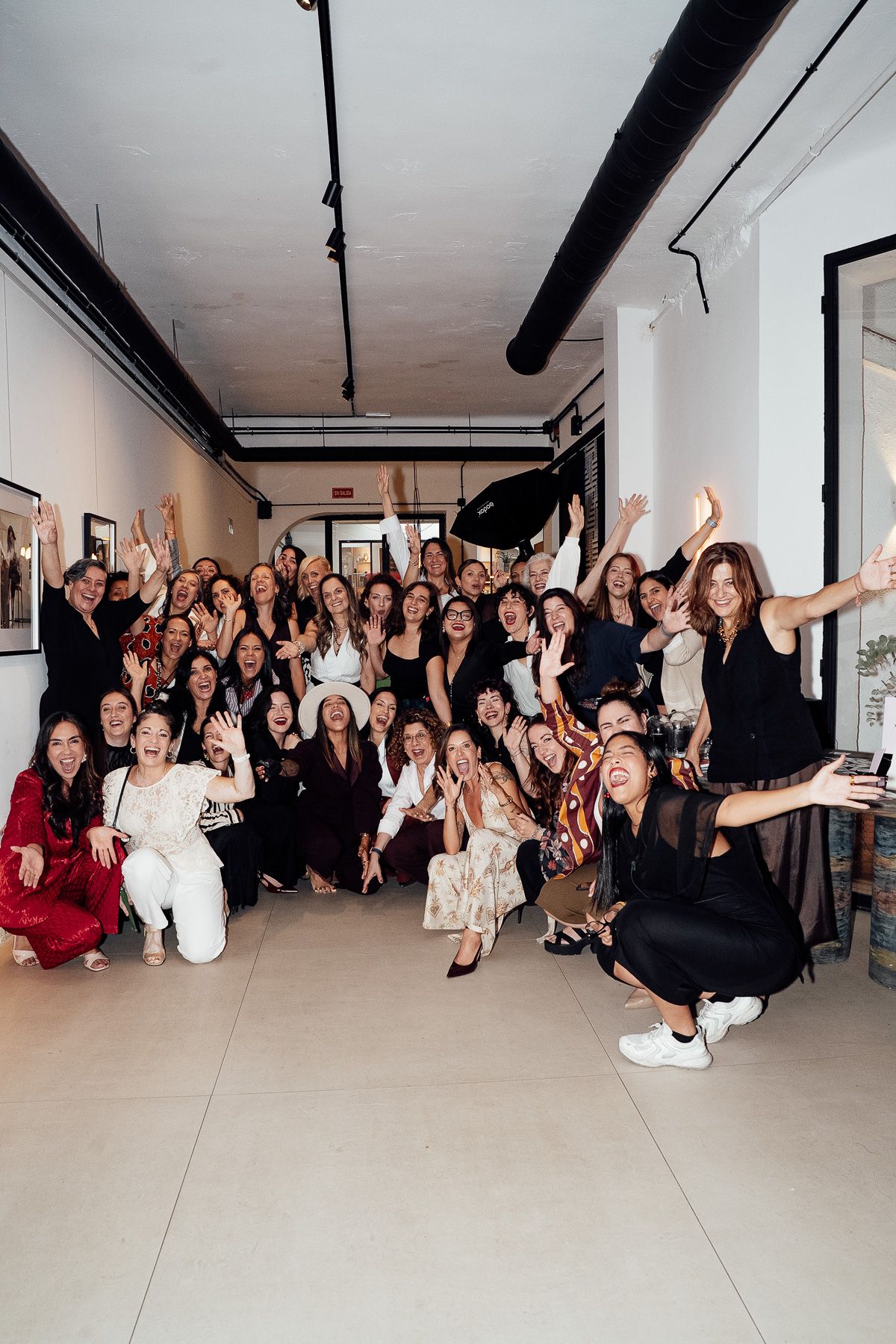 Large group of diverse women celebrating together in a modern white gallery space, arms raised in joy.