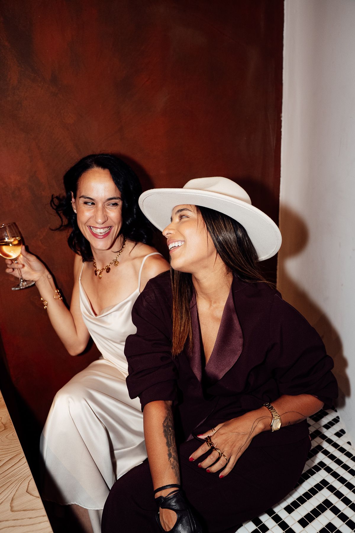 Two stylish women laughing together at an event, one in white satin dress, one in dark suit with hat.