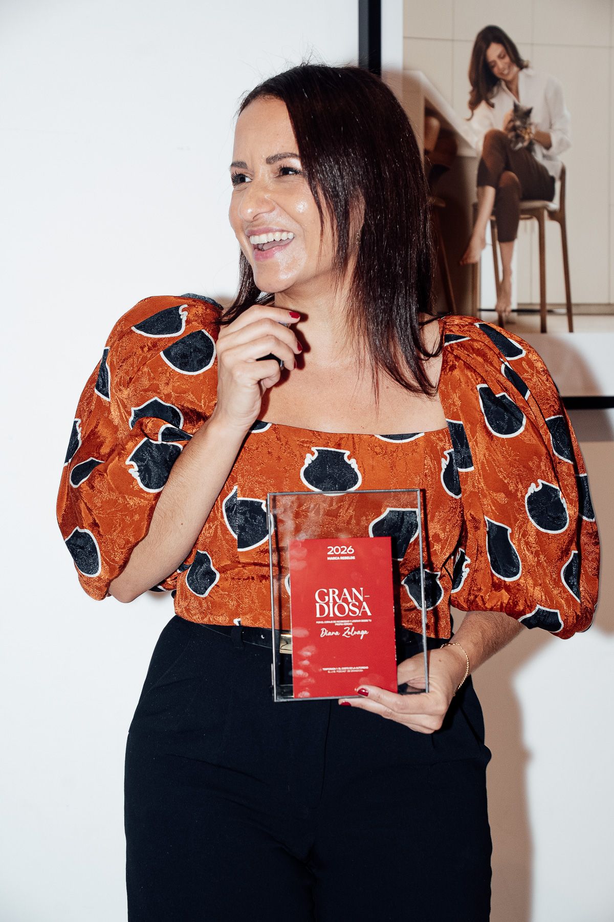 Woman in orange floral top smiling while holding a Gran Diosa award plaque at an indoor event.