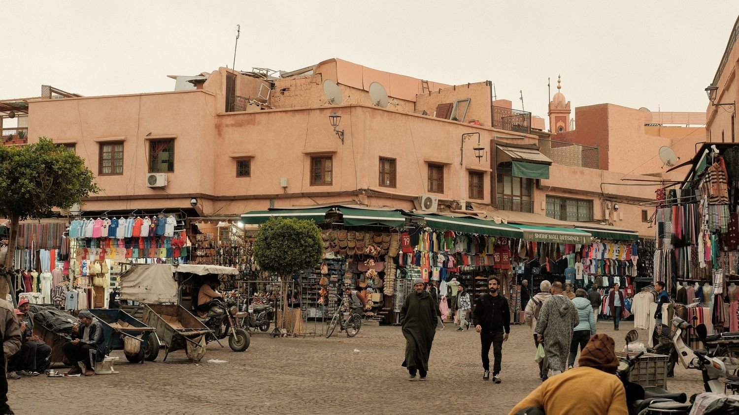 Bustling Marrakech medina market square with shops, pedestrians, and traditional pink buildings under overcast sky.