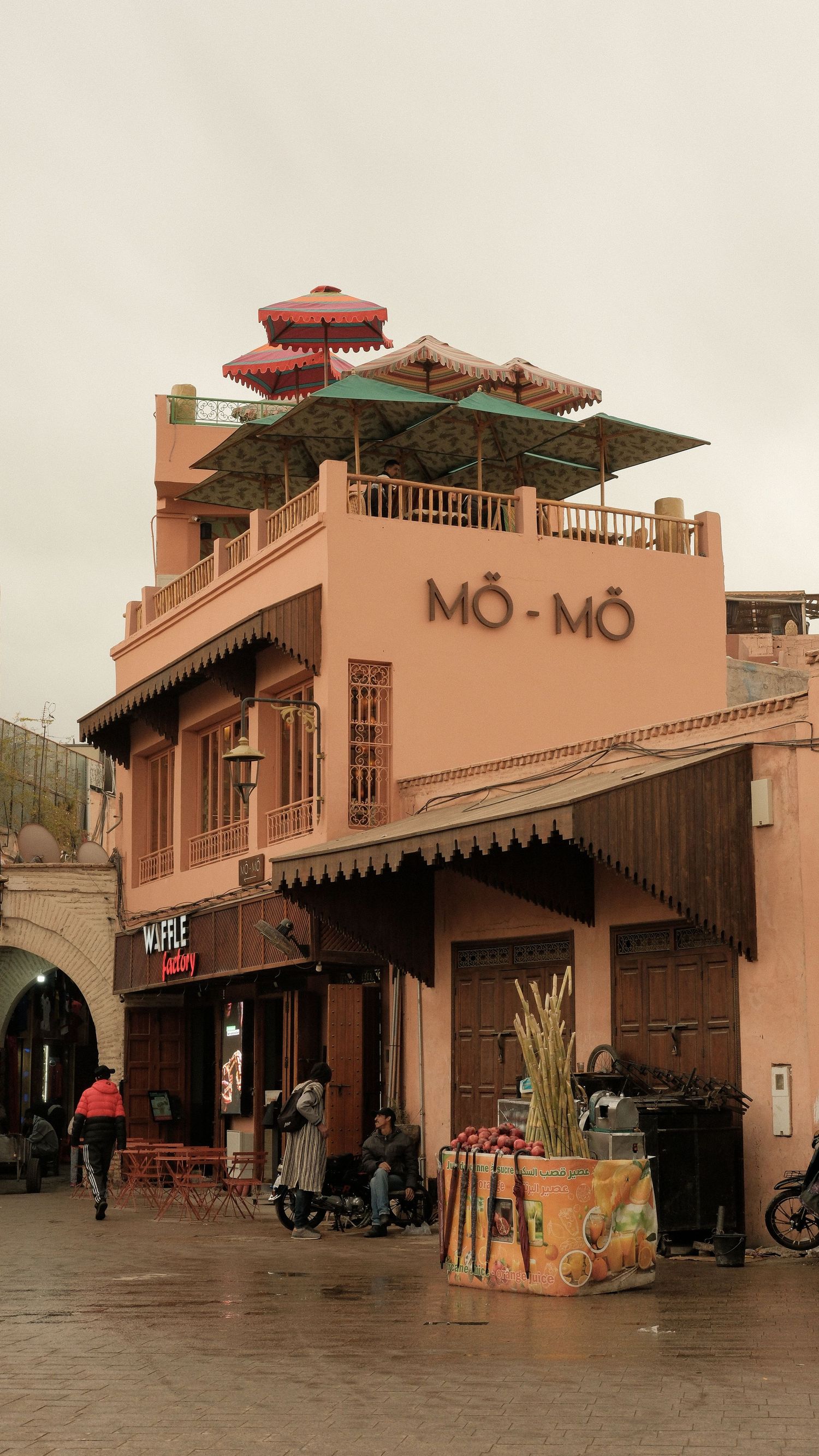 Pink Moroccan building with MO-MO restaurant sign, rooftop terrace with umbrellas, and Waffle shop nearby on a cloudy day.