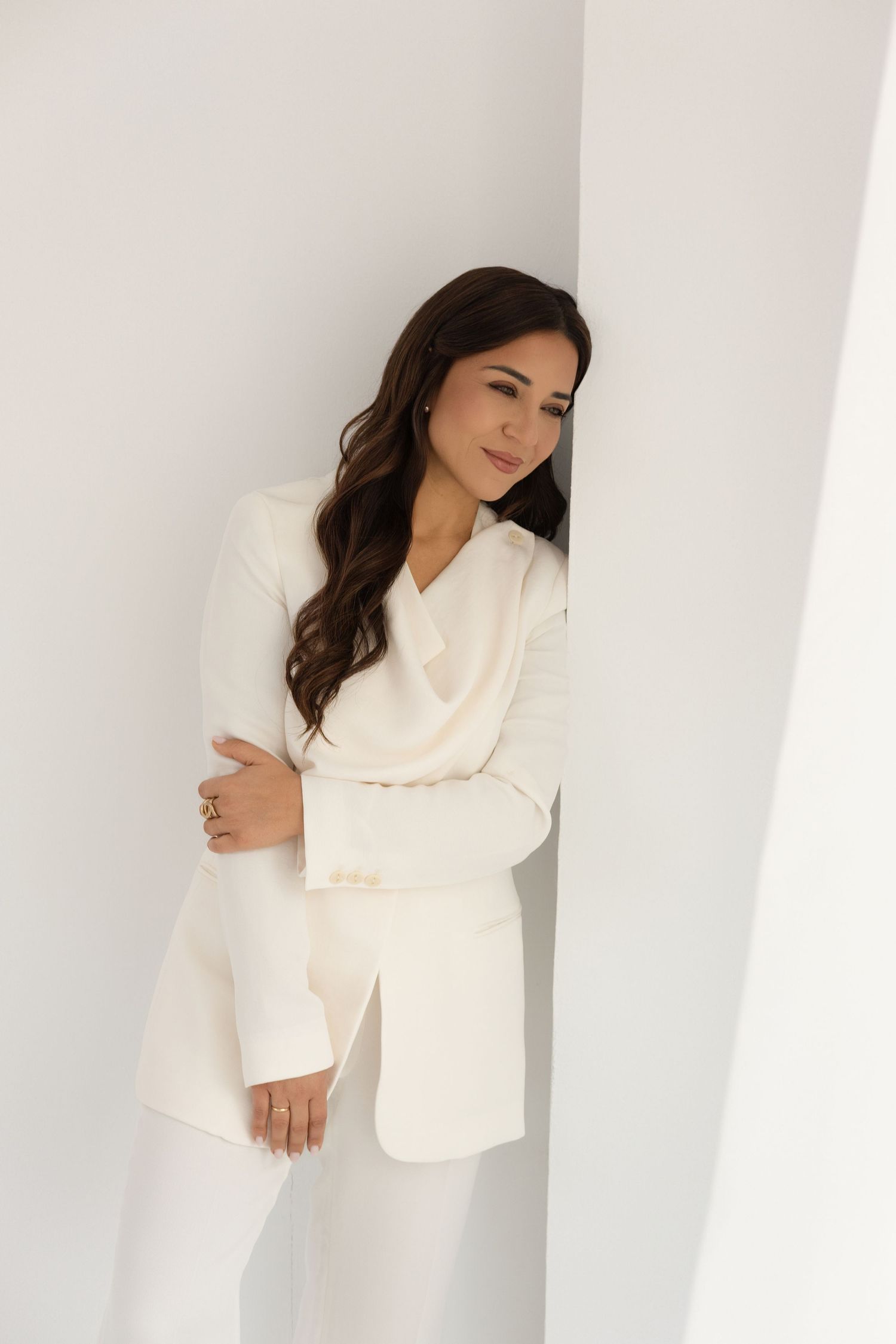 Woman in white blazer and pants leans against white wall, arms crossed, looking down elegantly.