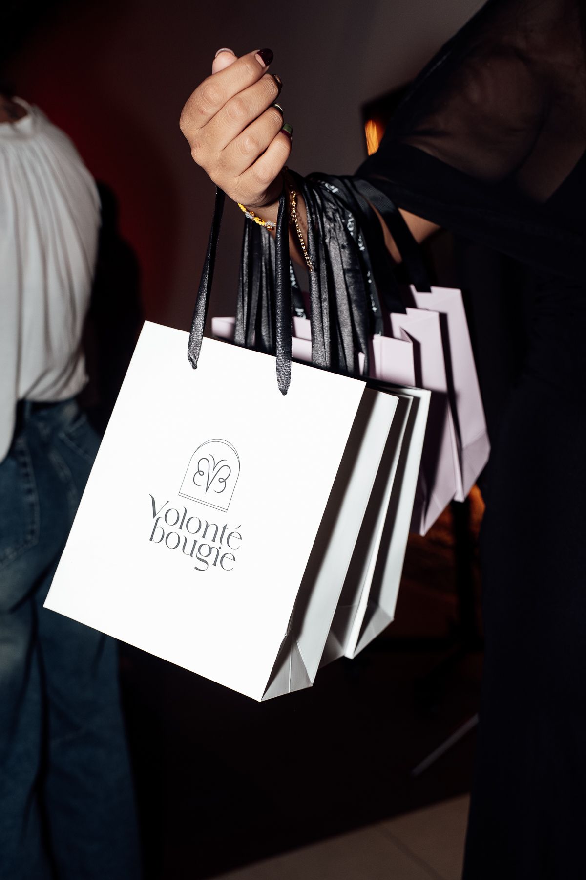 Hand holding multiple white Volonté Bougie luxury shopping bags with black handles at an event.