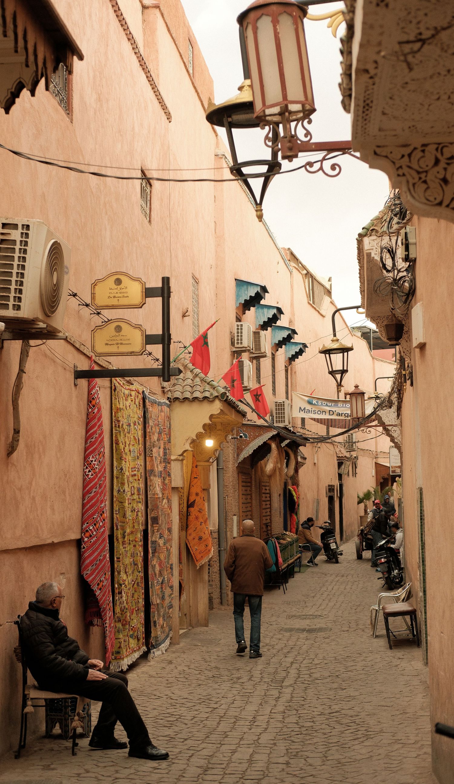 Narrow Moroccan medina alley with colorful rugs, lanterns, and pedestrians in Marrakech's historic souks.