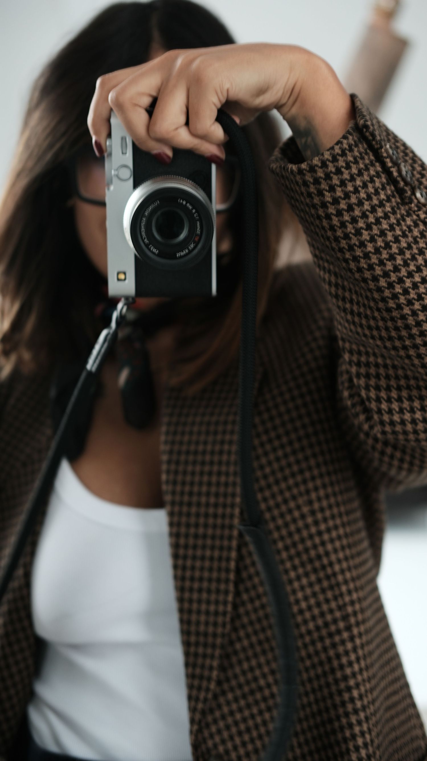 Woman in houndstooth blazer holding a compact camera up to her face against a white background.