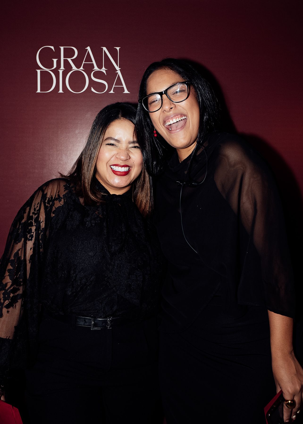 Two women laughing together in black outfits in front of Gran Diosa branded backdrop.