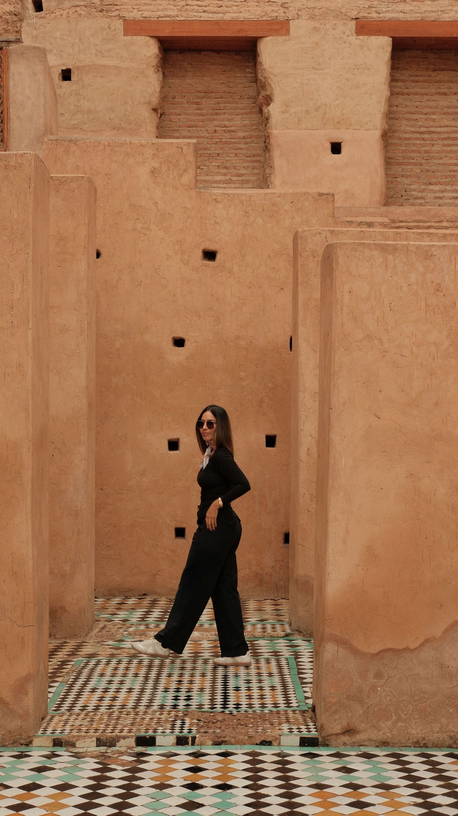 Woman in black outfit posing at ancient Moroccan clay architecture with colorful mosaic tile floor.