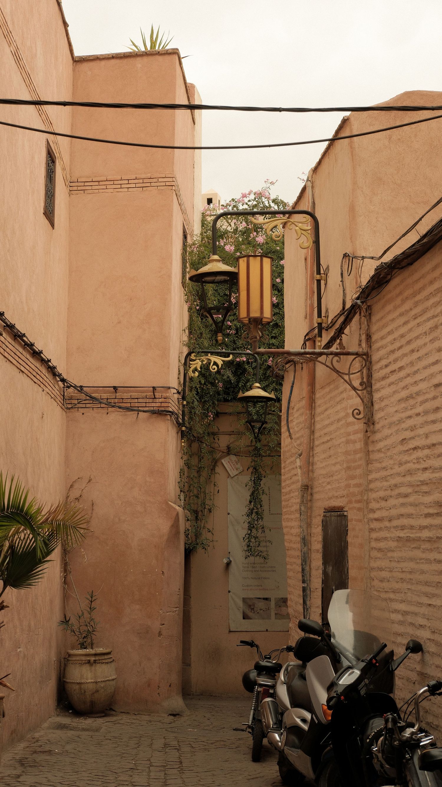 Narrow alleyway in Marrakech medina with pink walls, lanterns, ivy, parked motorcycles, and potted plants.