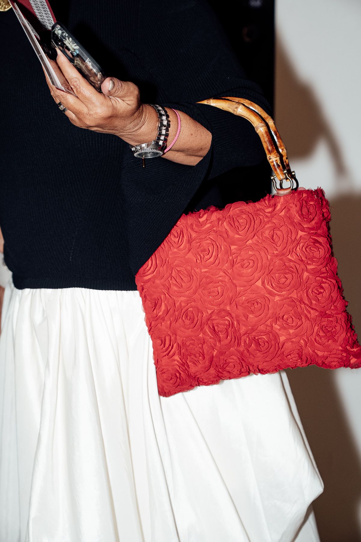 Woman holding red rose-textured clutch bag with bamboo handle, wearing white skirt and black top.