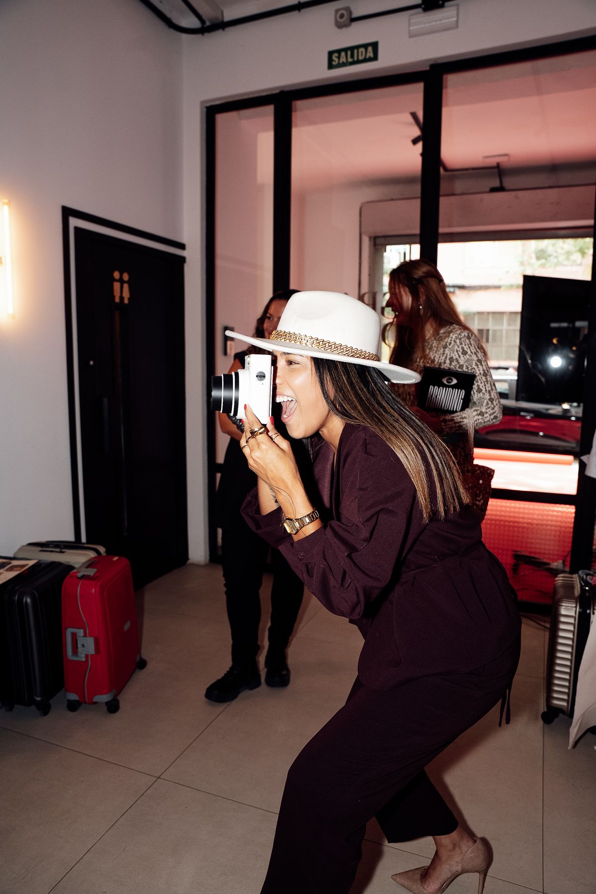 Woman in white hat and burgundy outfit crouches to take photo with camera at indoor event.