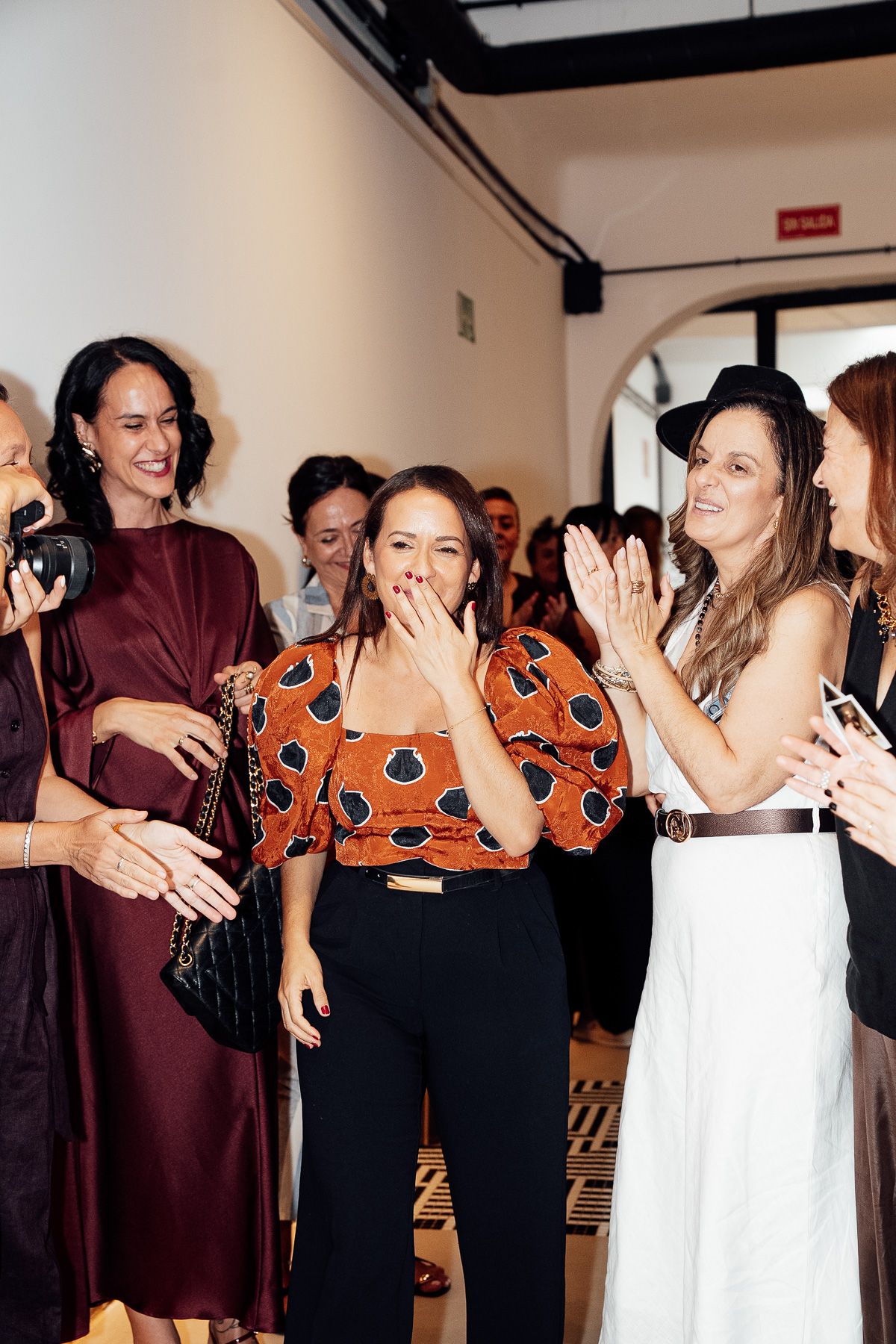 A group of stylish women celebrating together at an indoor event, with one woman in an orange top covering her mouth in surprise.