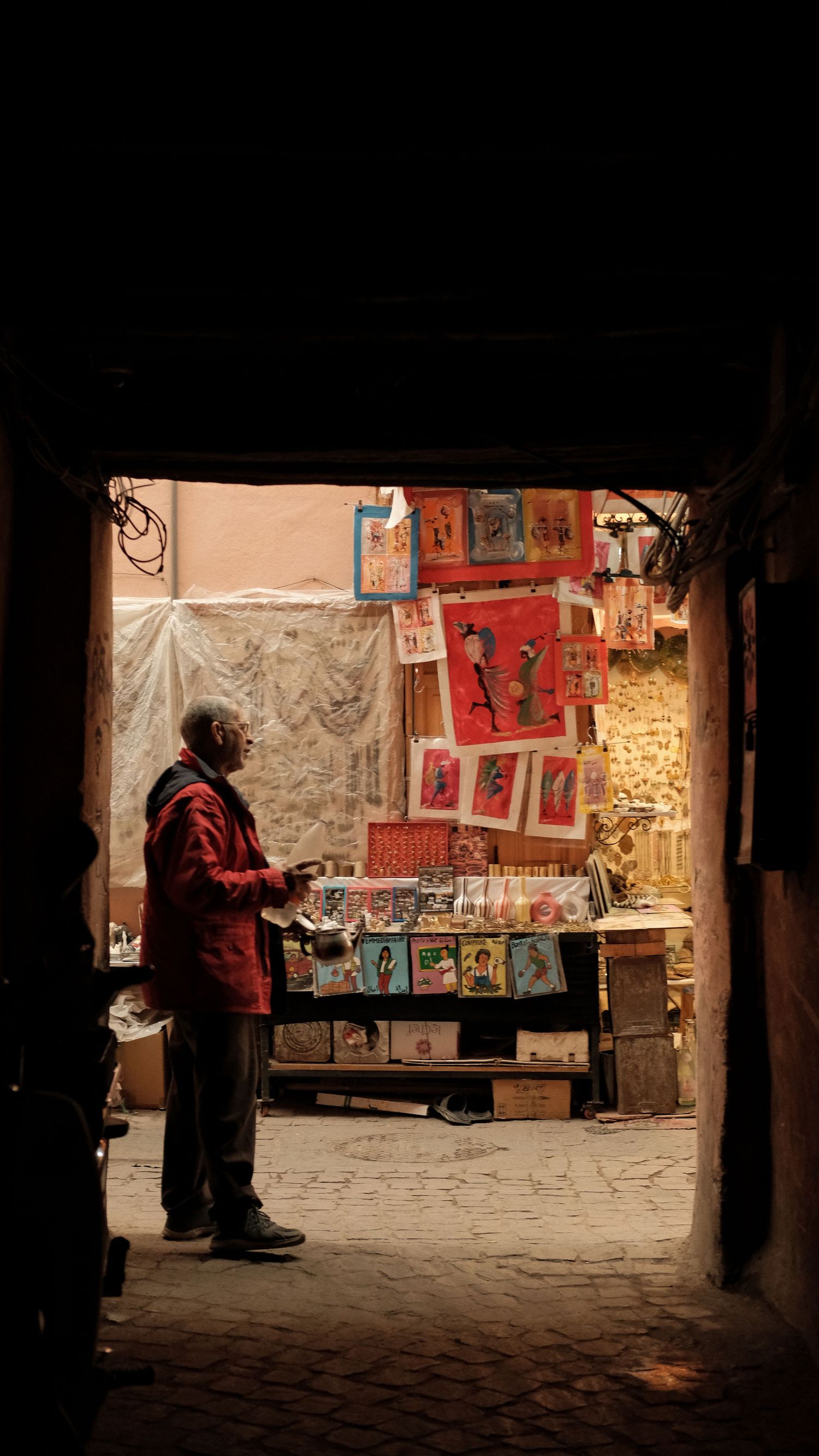 Man in red jacket browsing colorful art prints at a dimly lit outdoor market stall in a narrow alley.