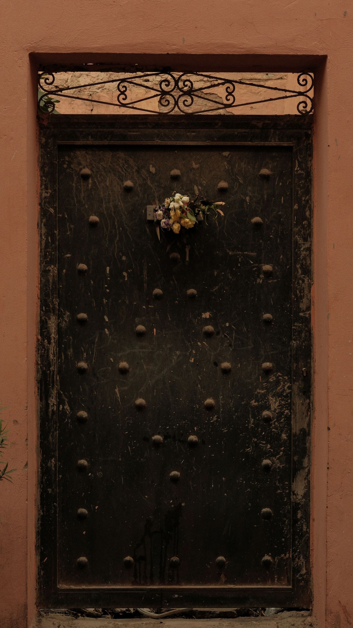Ancient studded wooden door with ornate iron scrollwork transom and small floral decoration on a terracotta wall.