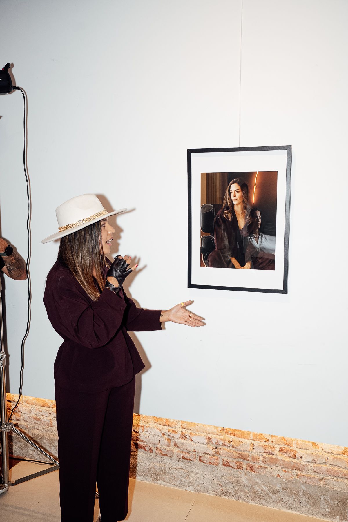 Woman in white hat and burgundy suit gesturing toward framed portrait photograph on gallery wall.