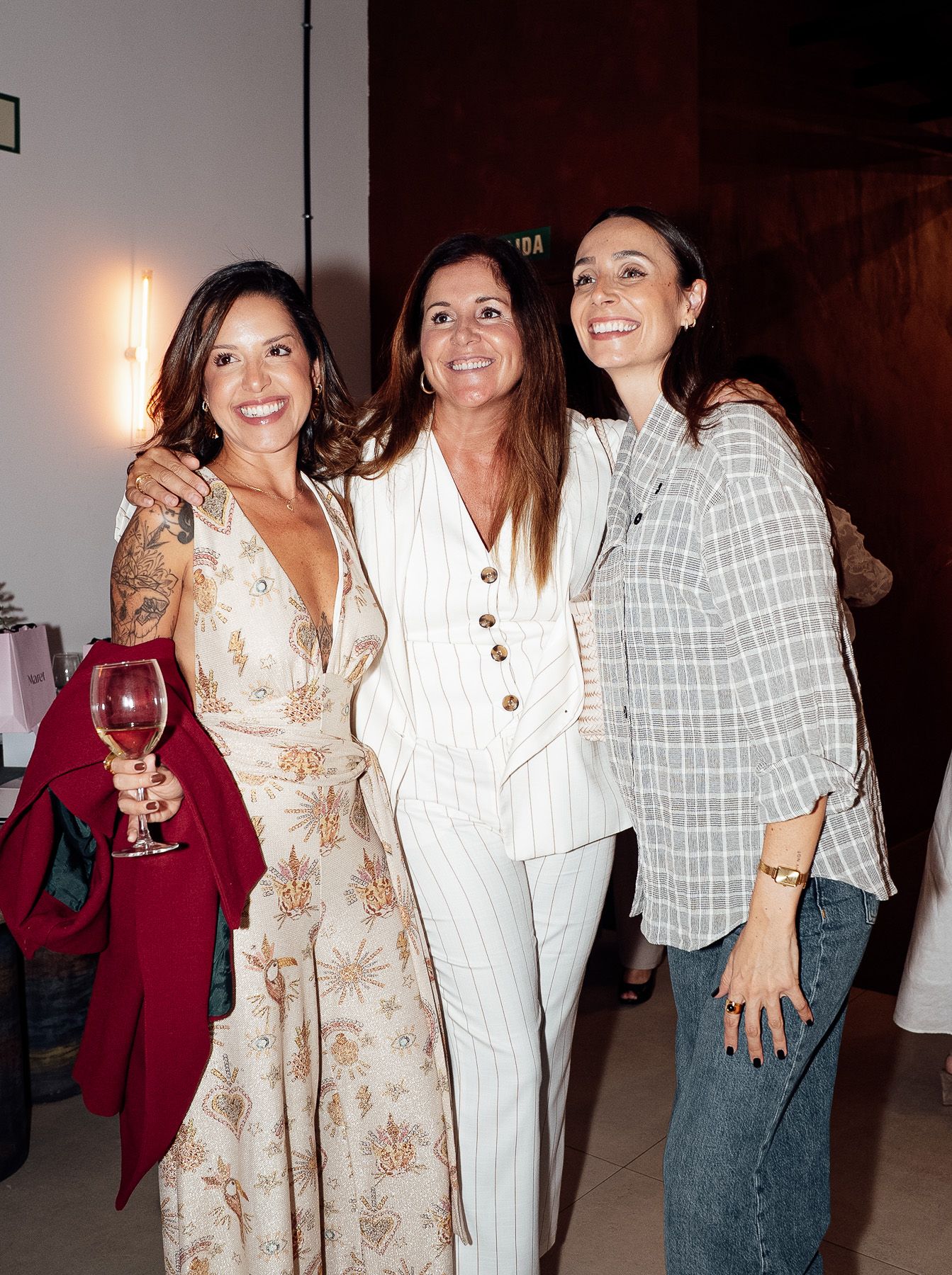 Three women smiling and posing together at a social gathering, one holding a wine glass.