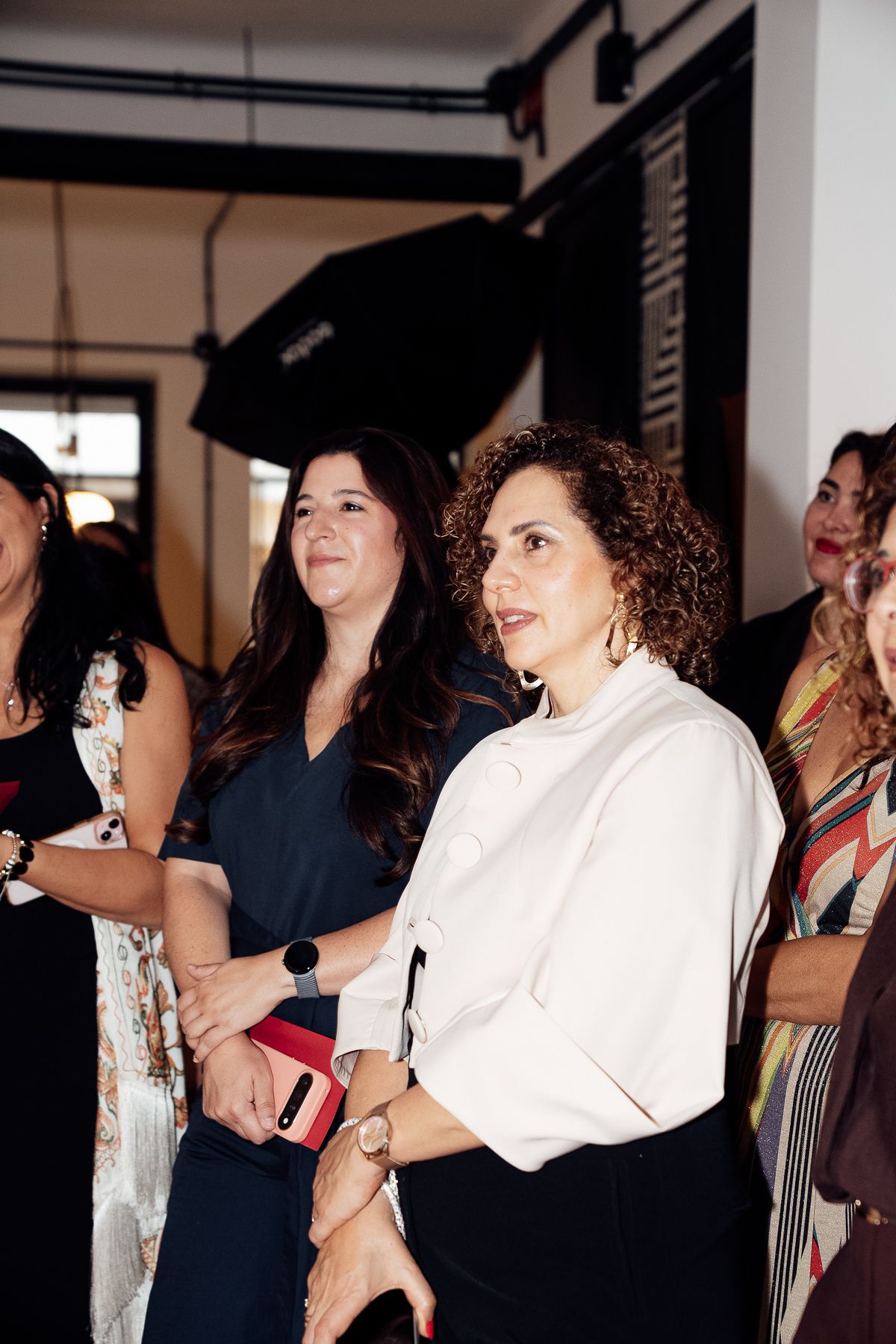 Women in elegant attire attentively watching an event at a modern indoor venue.